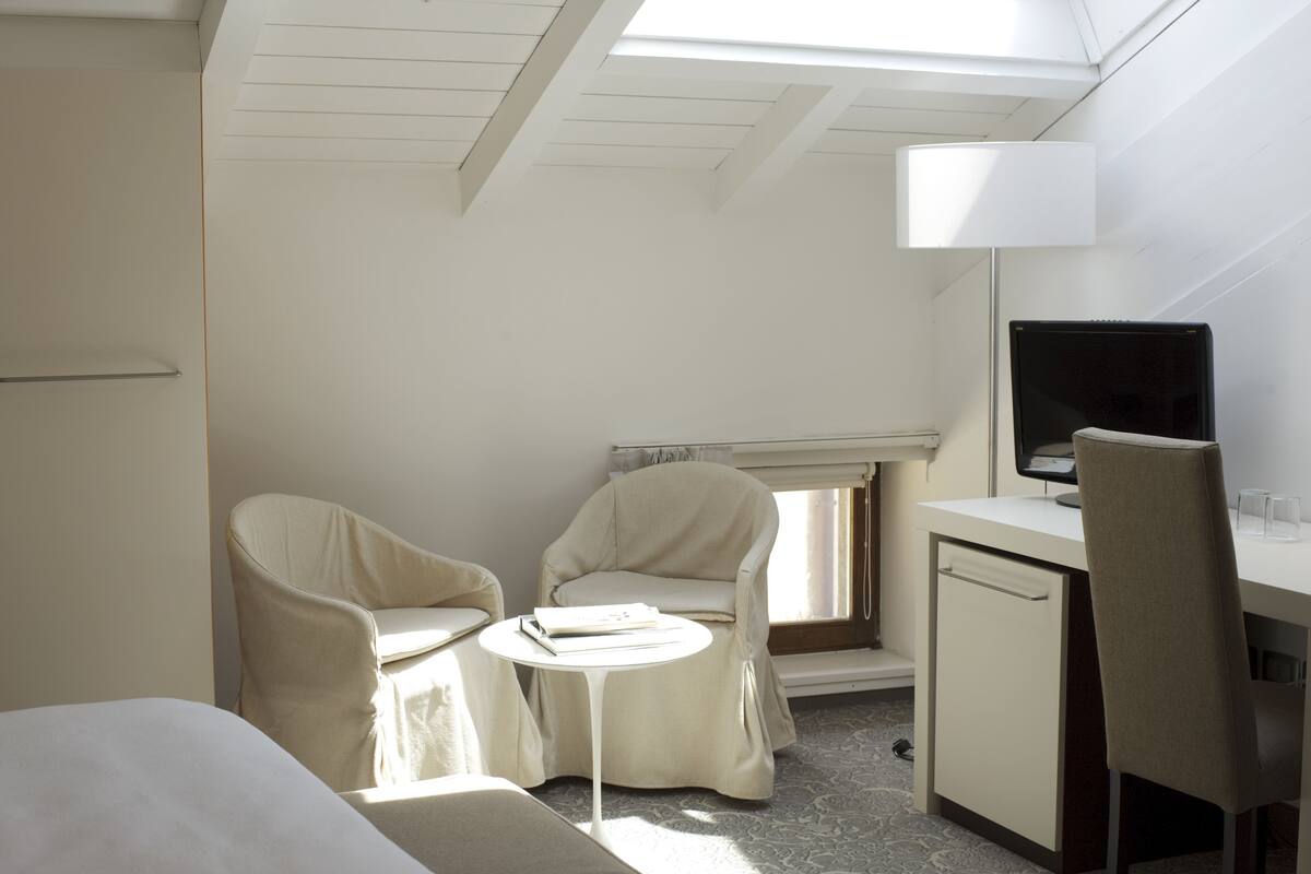 Une chambre d’hôtel comprenant un lit, deux chaises, une petite table, un bureau avec un ordinateur et une fenêtre avec des stores.