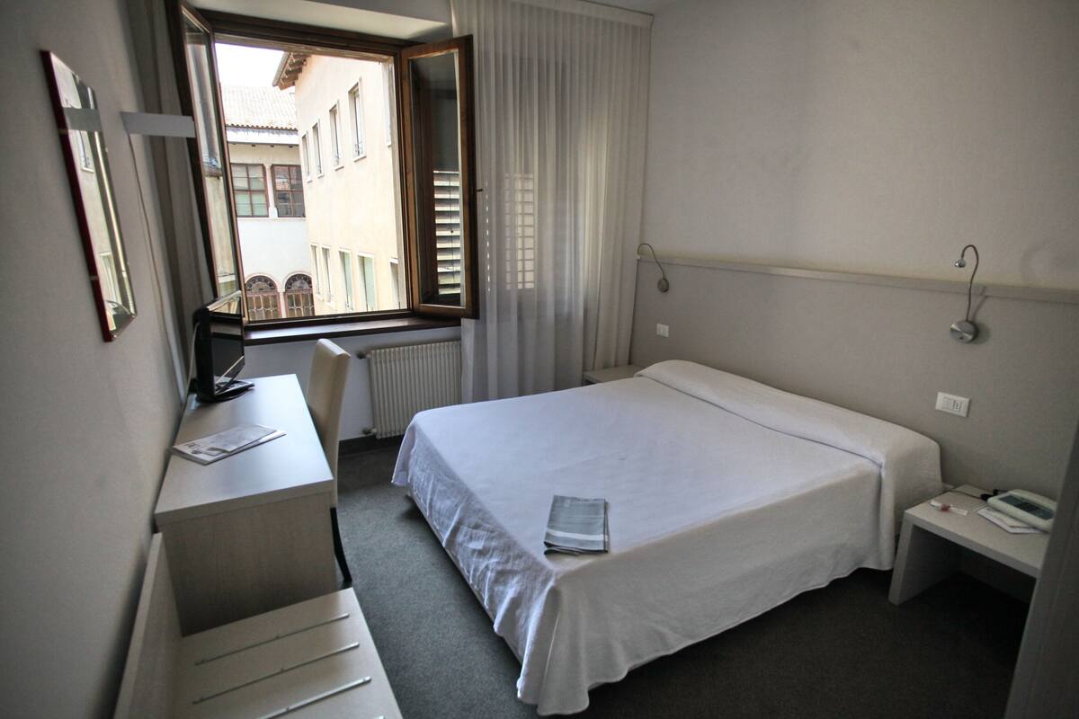 Une chambre d’hôtel comprenant un lit, un bureau, une chaise, une fenêtre avec des rideaux et une vue sur les bâtiments environnants.