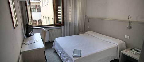 Habitación de hotel con cama, escritorio, silla, ventana con cortinas y vista a edificios.