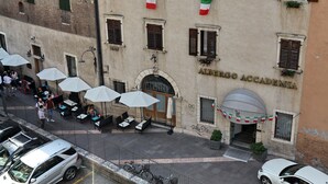 Een terras met parasols, een gebouw met het bordje "ALBERGO ACCADEMIA", en een straat met geparkeerde auto's en fietsen.