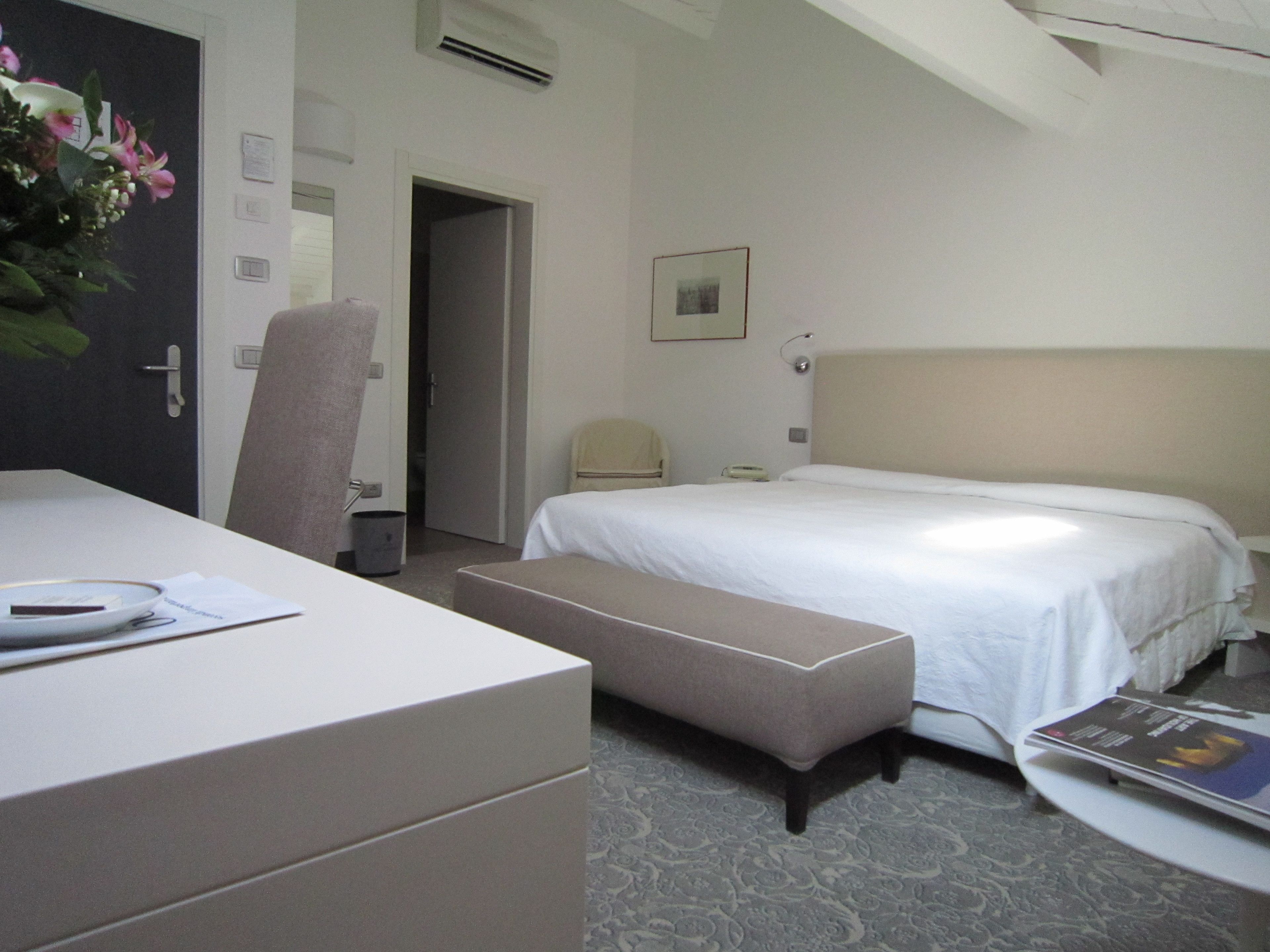 Quarto de hotel com cama, escrivaninha, cadeira, mesa de cabeceira e uma pequena mesa com uma revista.