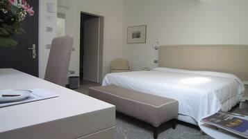 Quarto de hotel com cama, escrivaninha, cadeira, mesa de cabeceira e uma pequena mesa com uma revista.