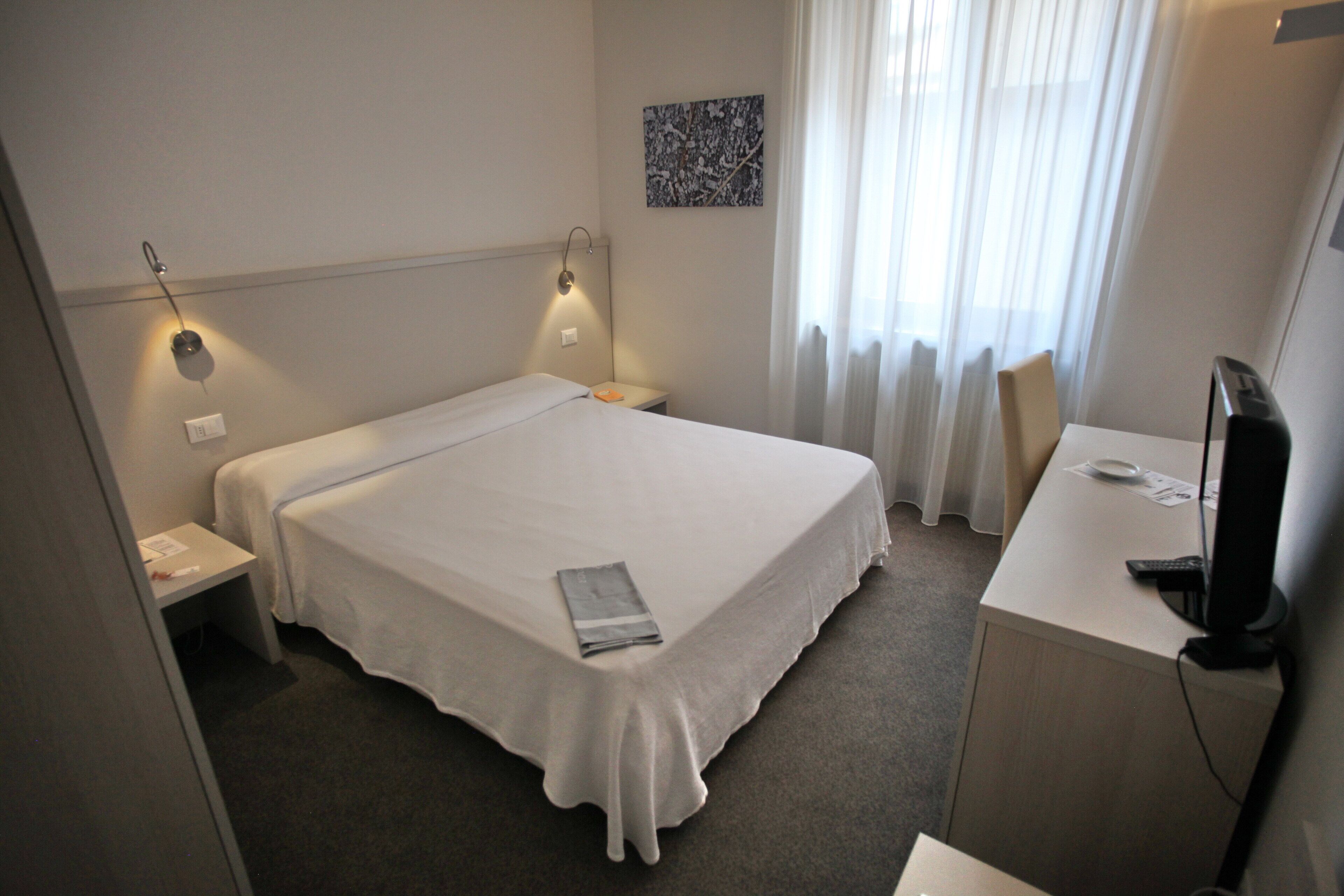 Ein Hotelzimmer mit Bett, Nachttischen, einem Schreibtisch mit Computer und einem Fenster mit Vorhängen.