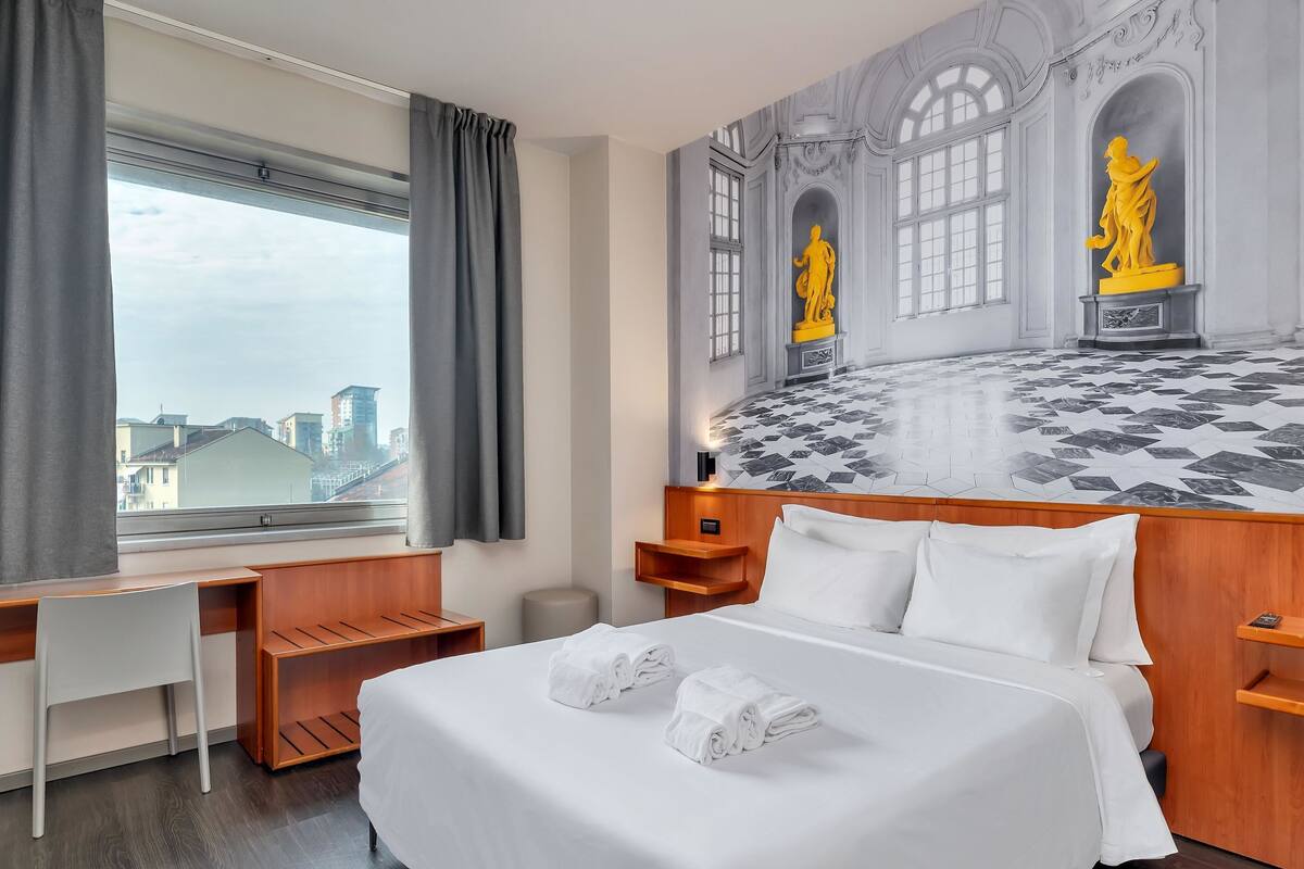 A hotel room with a bed, a desk, and a view of the city.