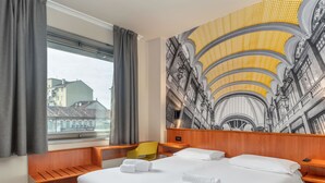 Habitación de hotel con una cama grande, cabecera de madera, un pequeño banco de madera, una silla amarilla y un amplio ventanal con cortinas.