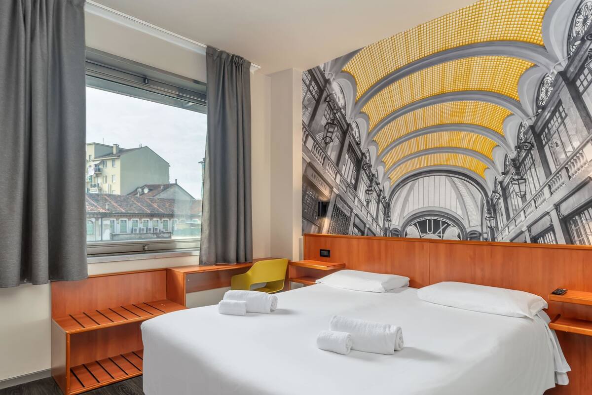 A hotel room with a large bed, a wooden headboard, a small wooden bench, a yellow chair, and a large window with curtains.