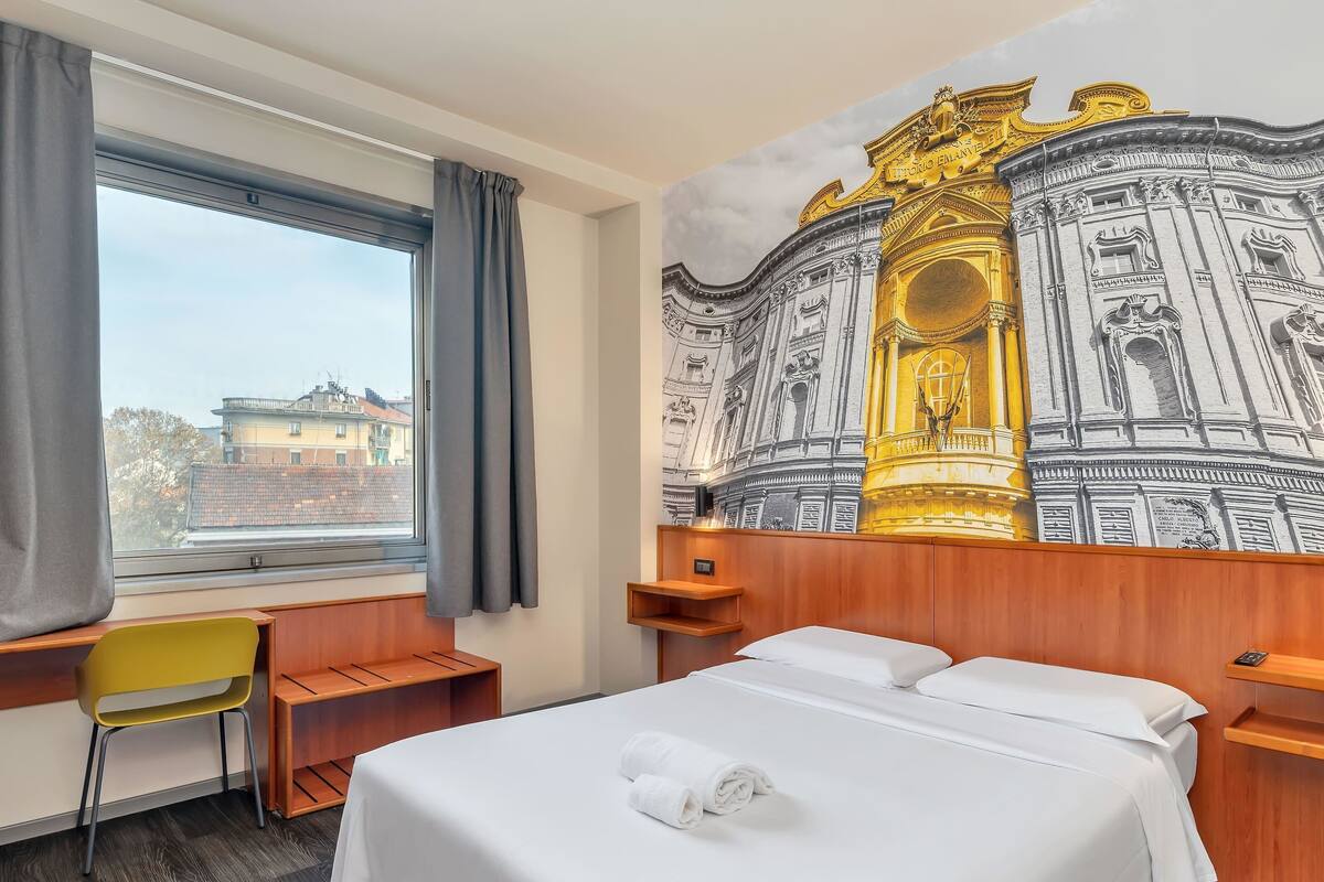 A hotel room with a bed, a desk, a chair, and a large mural of a historical building.