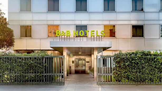 Entrada de un hotel con fachada moderna, un cartel con el nombre y un camino de acceso con portones.