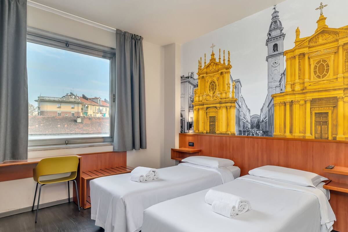 A hotel room with two beds, a wooden desk, a chair, and a large window.