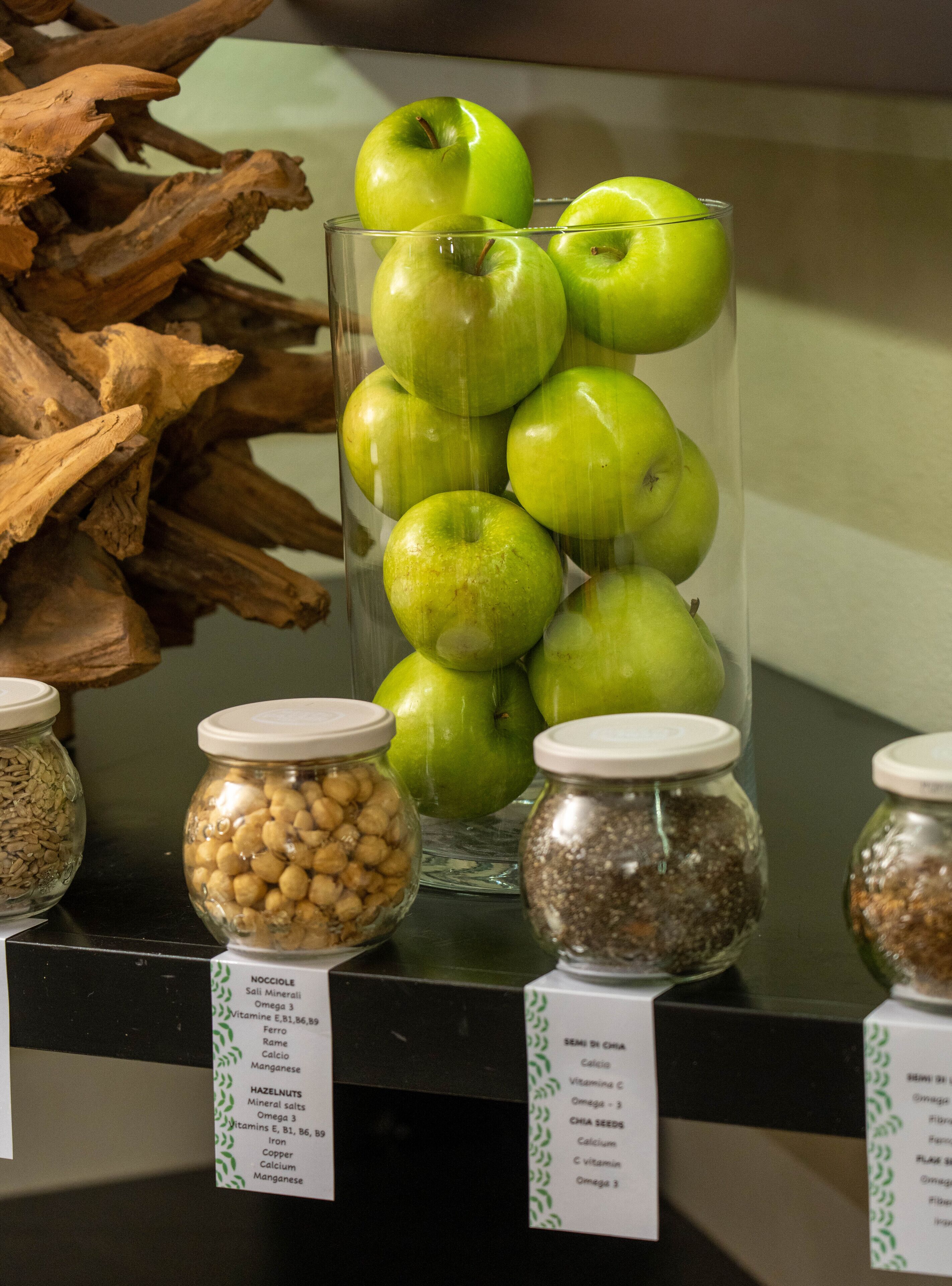 Een presentatie van groene appels in een glazen pot, omringd door potten met noten en zaden en voedingsinformatie labels.
