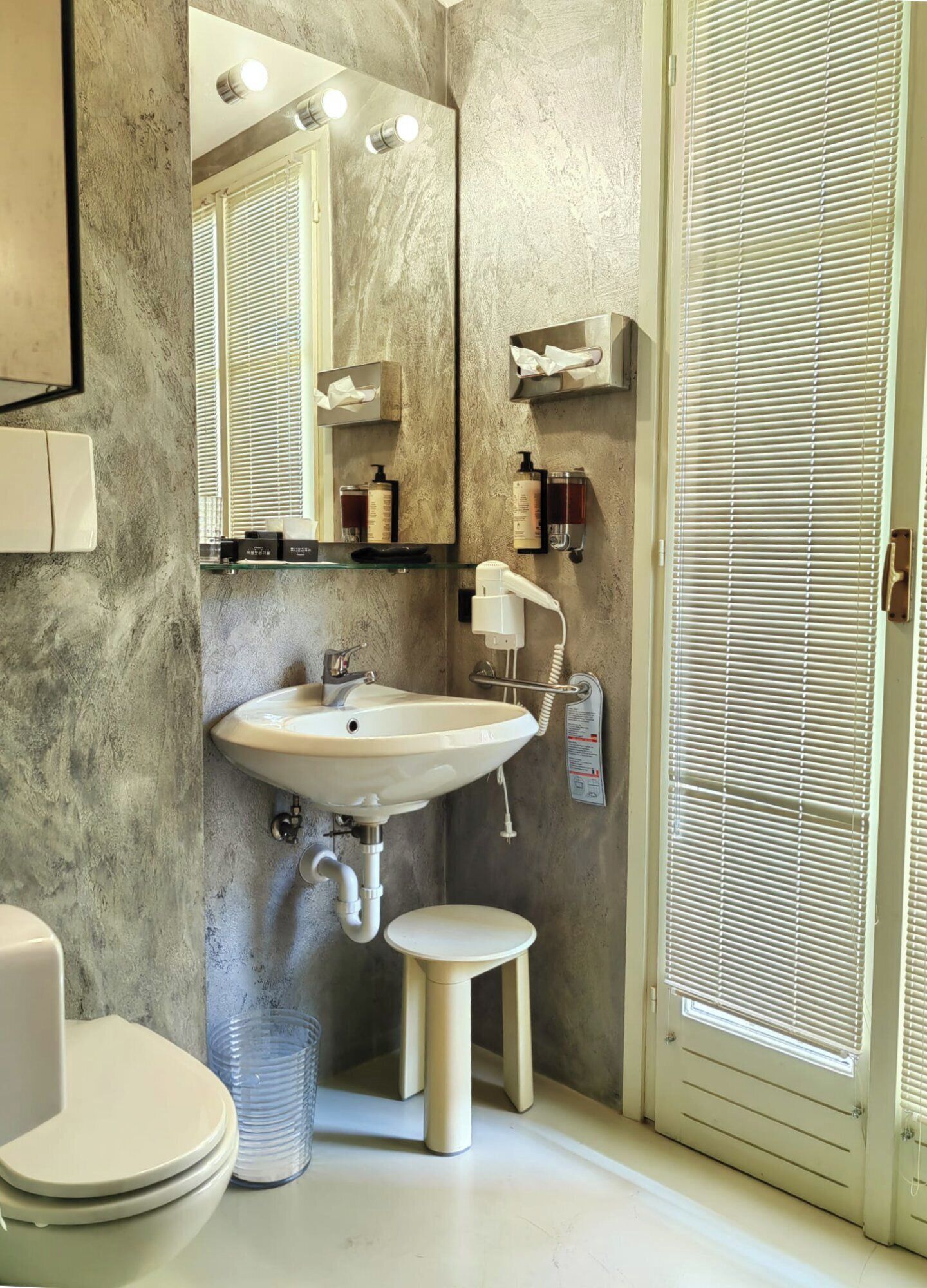 A bathroom with a white sink, a mirror, a toilet, and a bidet. There are shelves with toiletries and a towel rack.