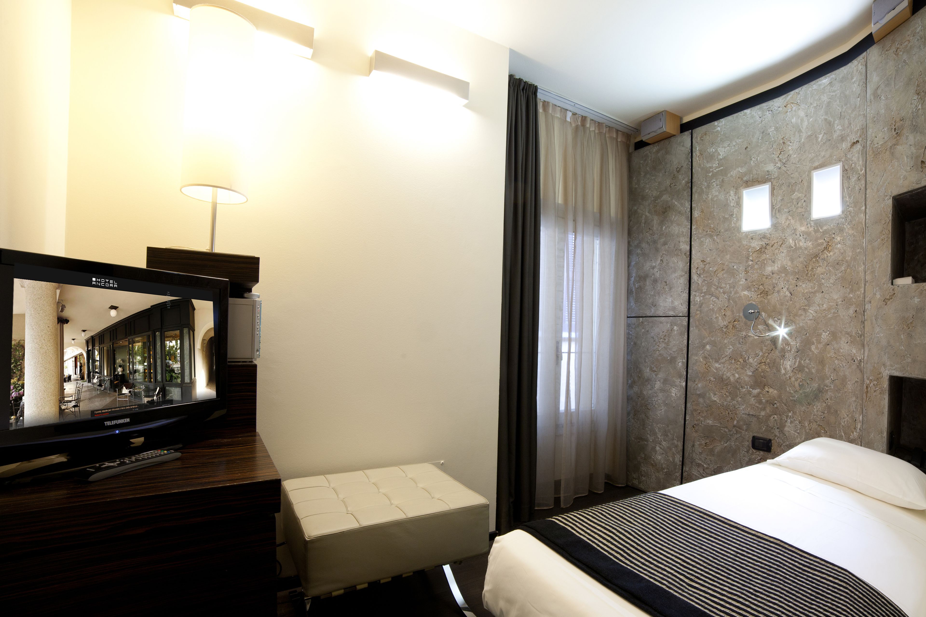 Una habitación de hotel con cama, mesita de noche con lámpara, televisión y un espejo grande.
