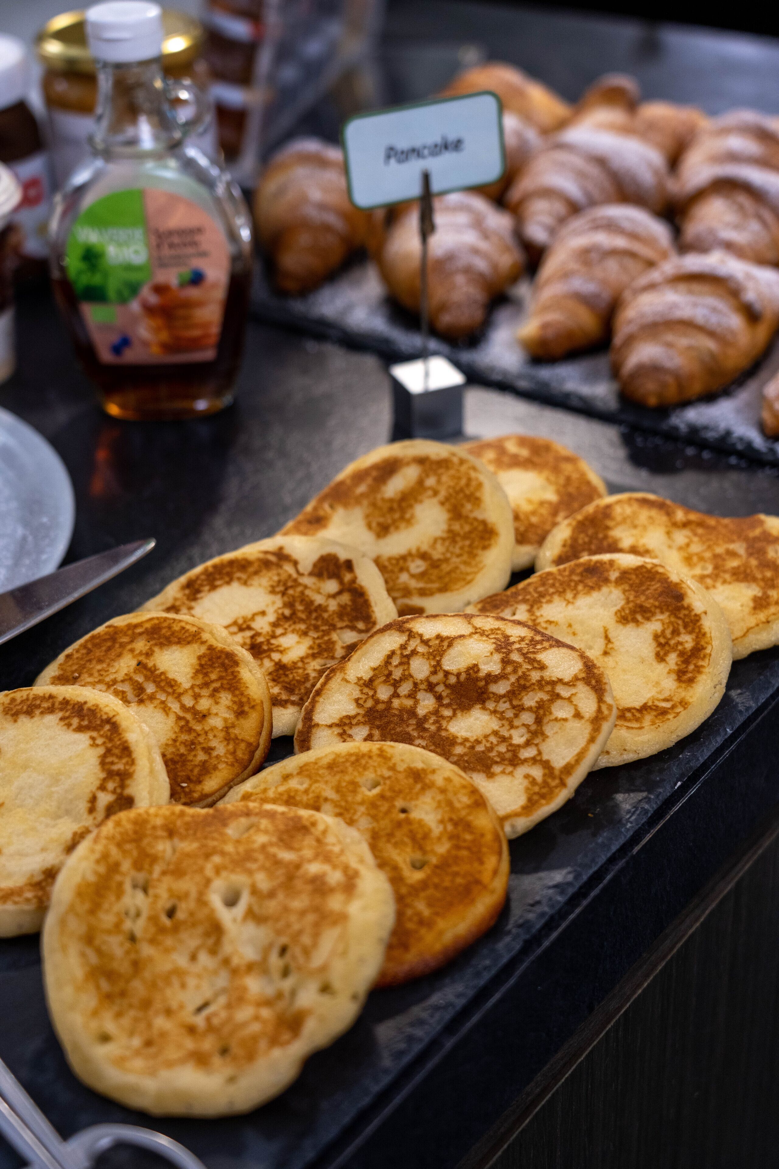 Un bufé de desayuno con panqueques, cruasanes y una botella de almíbar.
