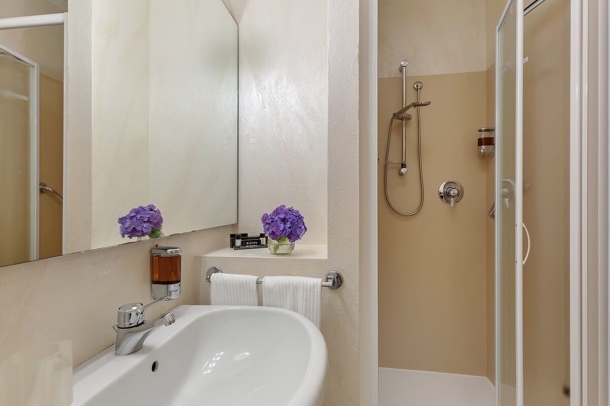 A bathroom with a white sink, a mirror, a shower, and a shelf with flowers.