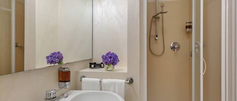 A bathroom with a white sink, a mirror, a shower, and a shelf with flowers.