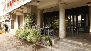 A hotel entrance with a covered outdoor seating area, columns, and a flower bed. - Hotel Ancora (Verbania)