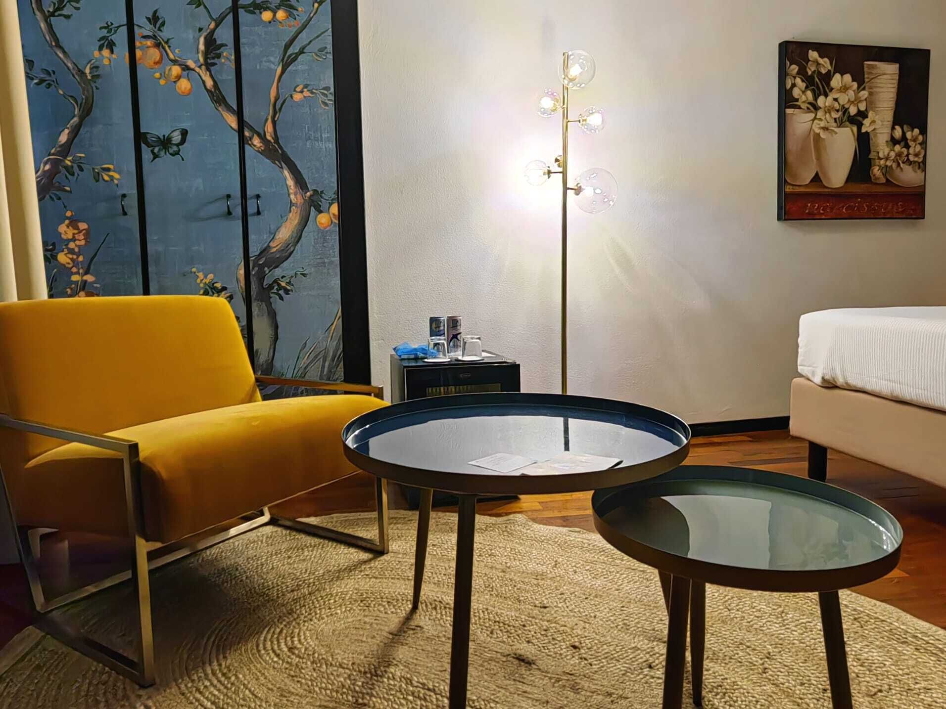 A modern living room with a yellow armchair, a glass-top coffee table, and a sofa. There is a painting on the wall and a floor lamp.