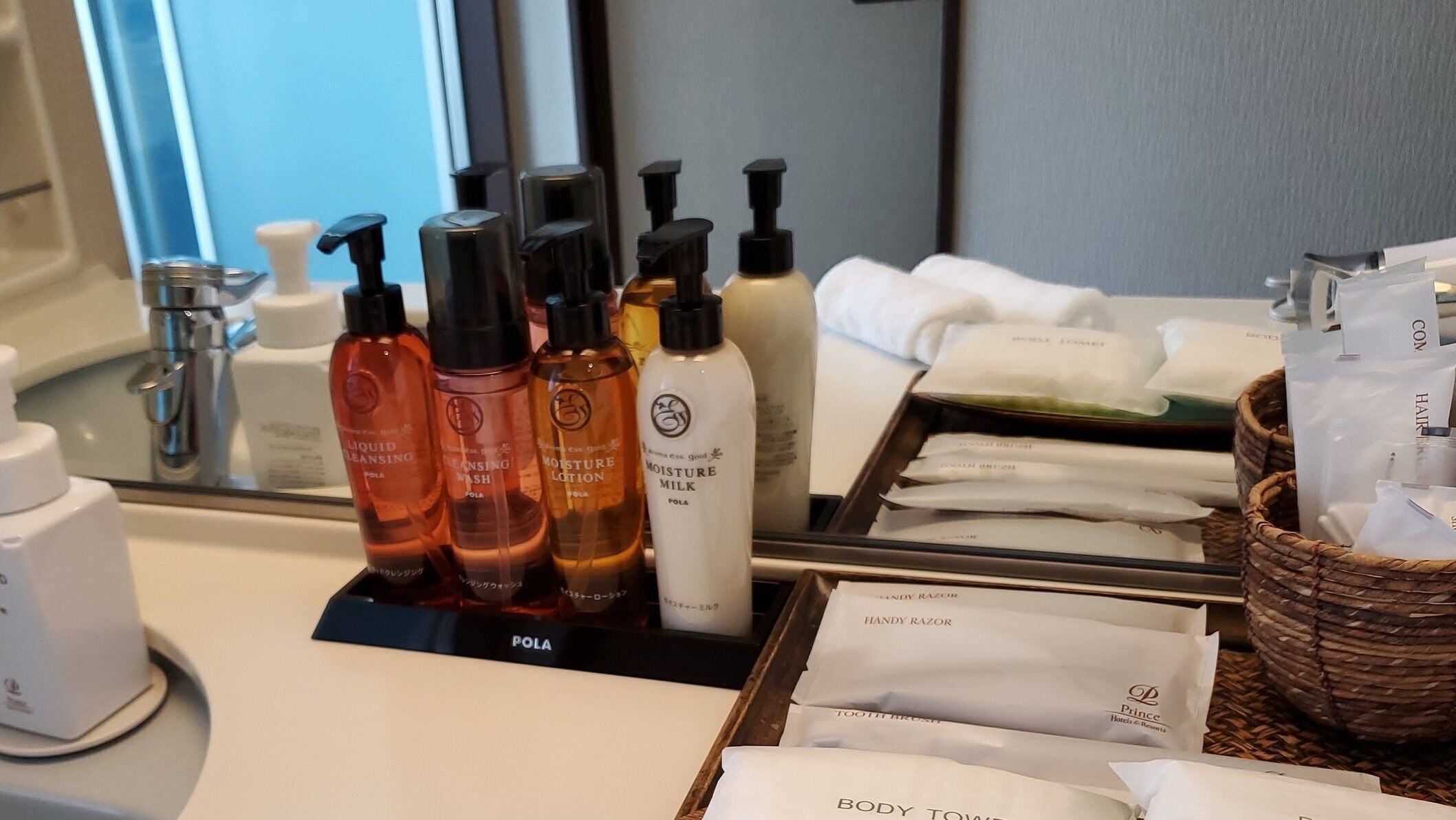 A bathroom counter with various toiletries including liquid cleansing, moisture lotion, and moisture milk. There are also white towels and a basket with additional items.