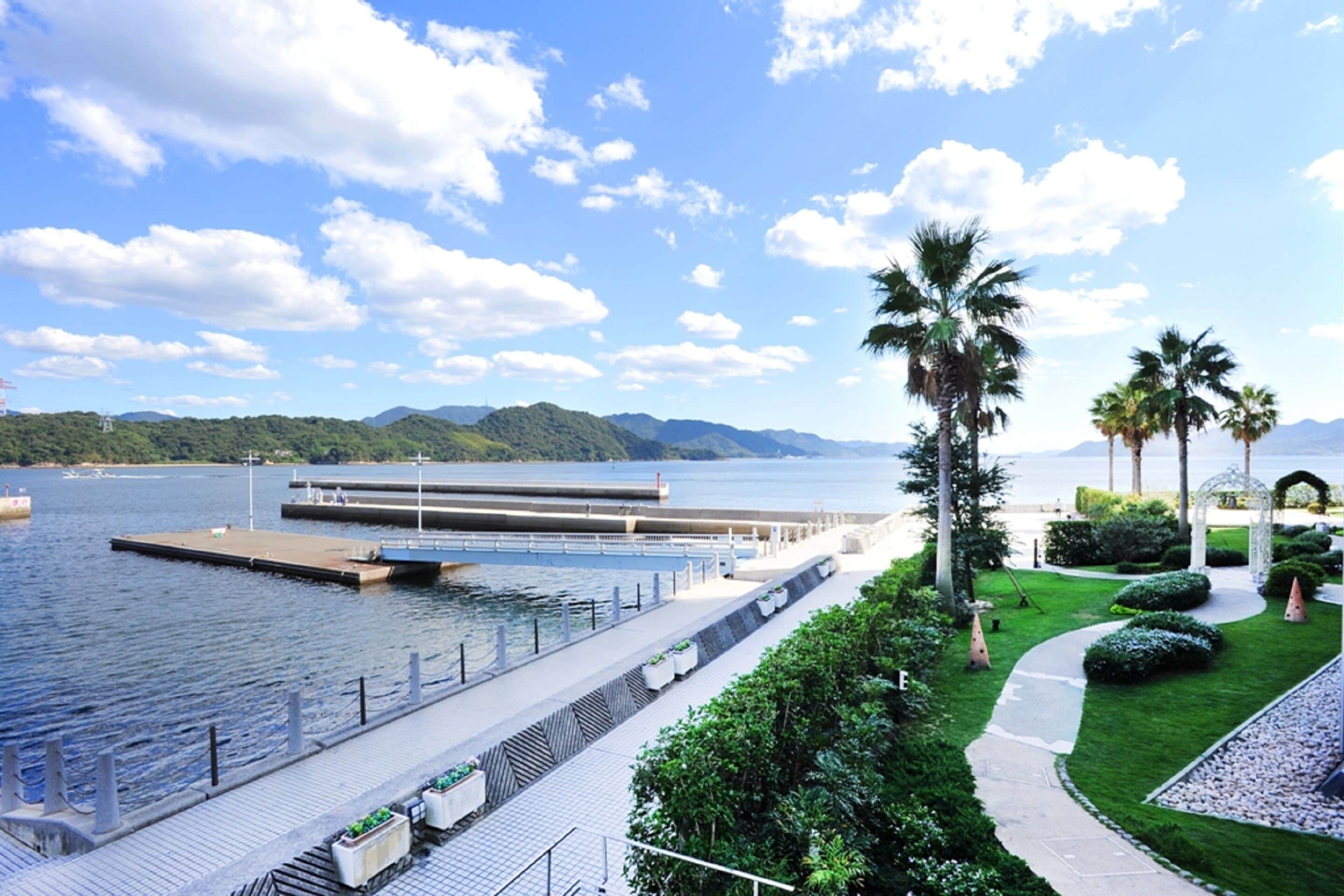 A waterfront promenade with a modern building, palm trees, and a clear sky.