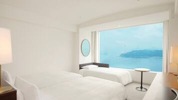 Quarto de hotel com duas camas, uma mesa redonda e uma janela ampla com vista para o mar.