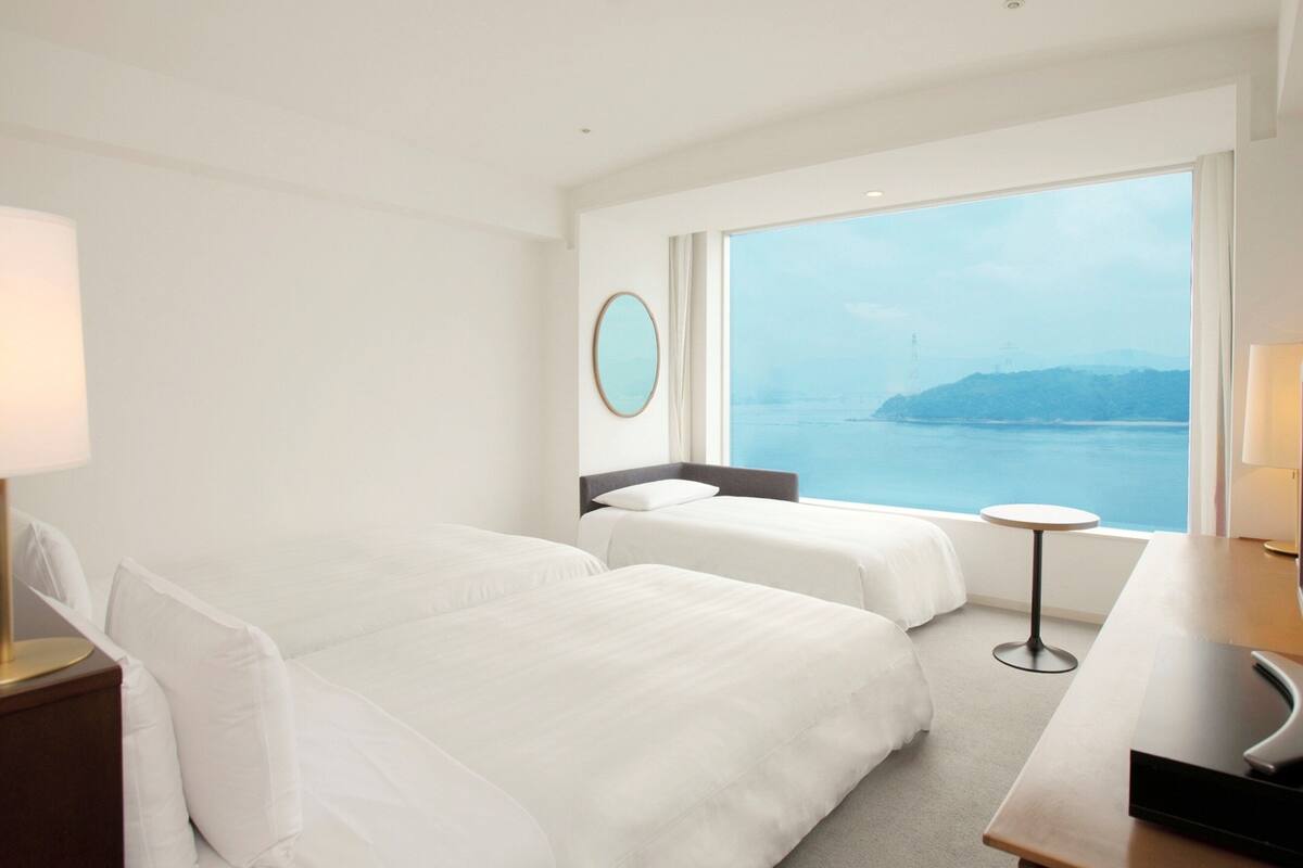 A hotel room with two beds, a round mirror, a small table, and a view of the sea.