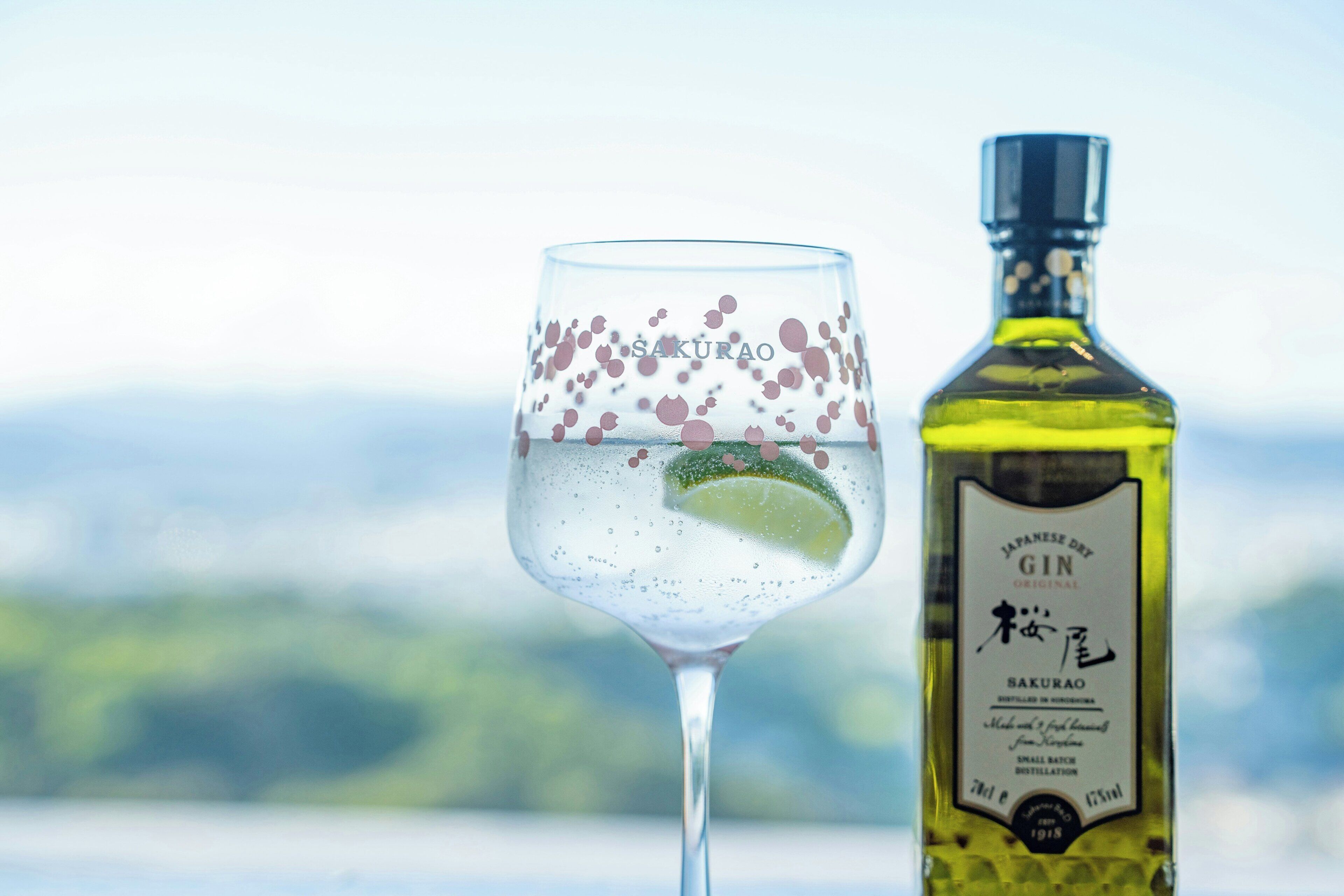 Et glas med et limeskive og bobler, ved siden af en flaske med teksten 'SAKURAO GIN'.