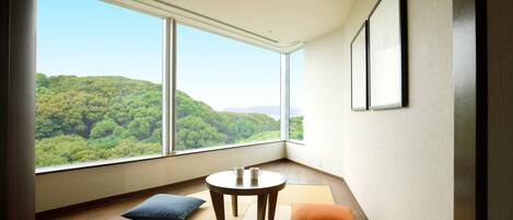 A room with a large window offering a view of a forested hill. There is a small round table with a pitcher and cups, and two pillows on the tatami mat floor.