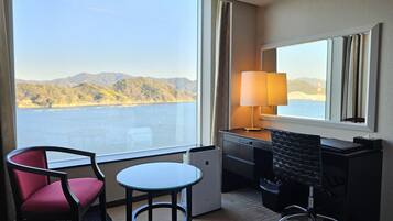 A hotel room with a large window offering a view of a body of water and hills, a desk with a lamp, a chair, and a small table.