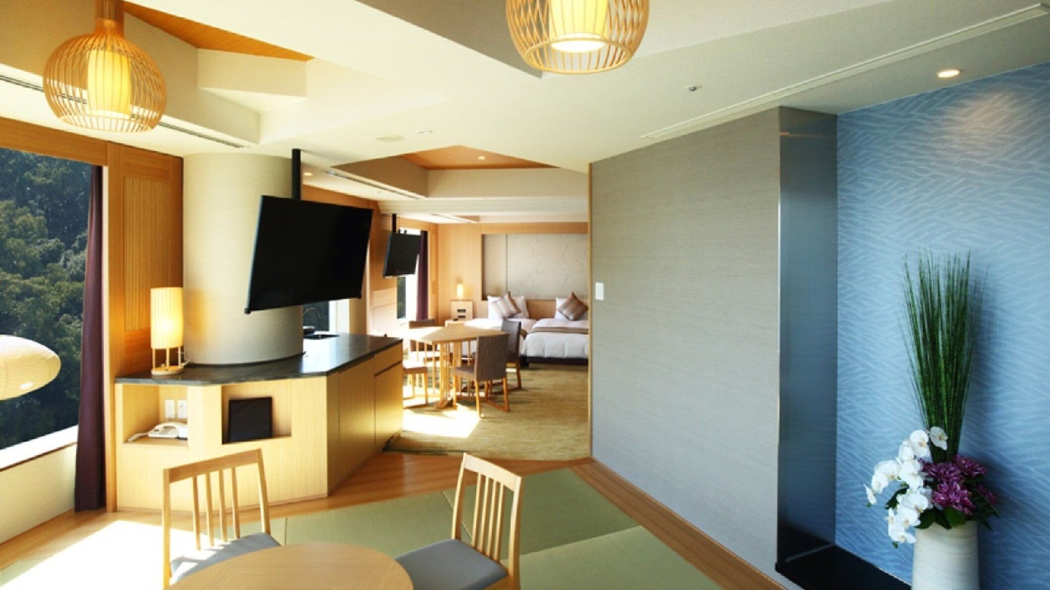 Une chambre d’hôtel moderne dotée d’un téléviseur à écran plat fixé sur une console en bois, d’un coin repas avec une table et des chaises, et d’une grande fenêtre offrant une vue sur l’extérieur.