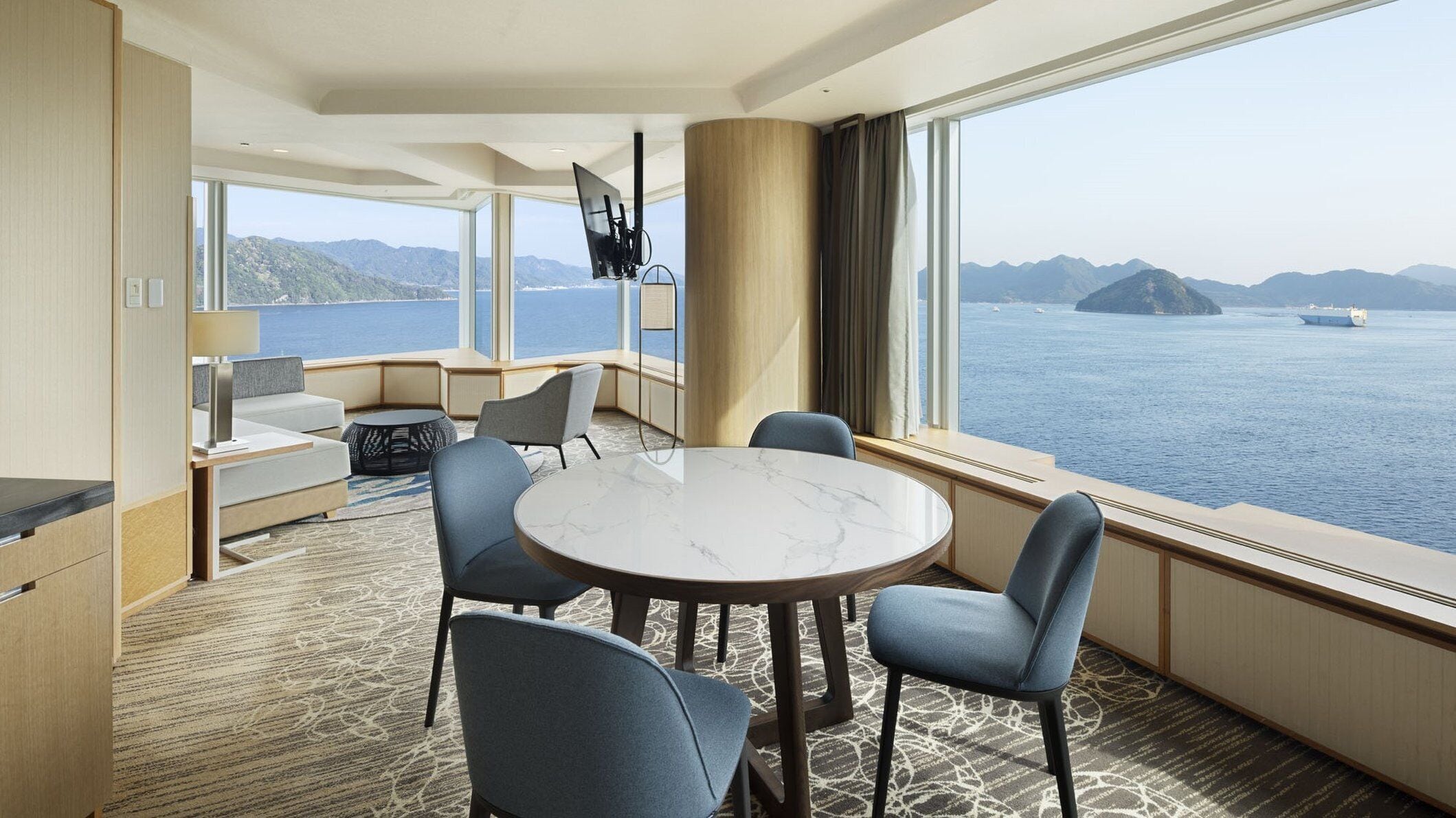 A modern hotel room with a round marble table, four chairs, a sofa, and a large window offering a view of the sea and mountains.