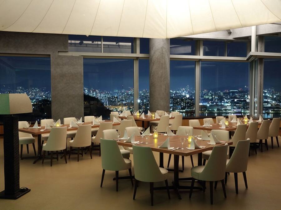 Comedor con varias mesas y sillas, ventanas amplias con vistas a la ciudad y un diseño moderno de techo.
