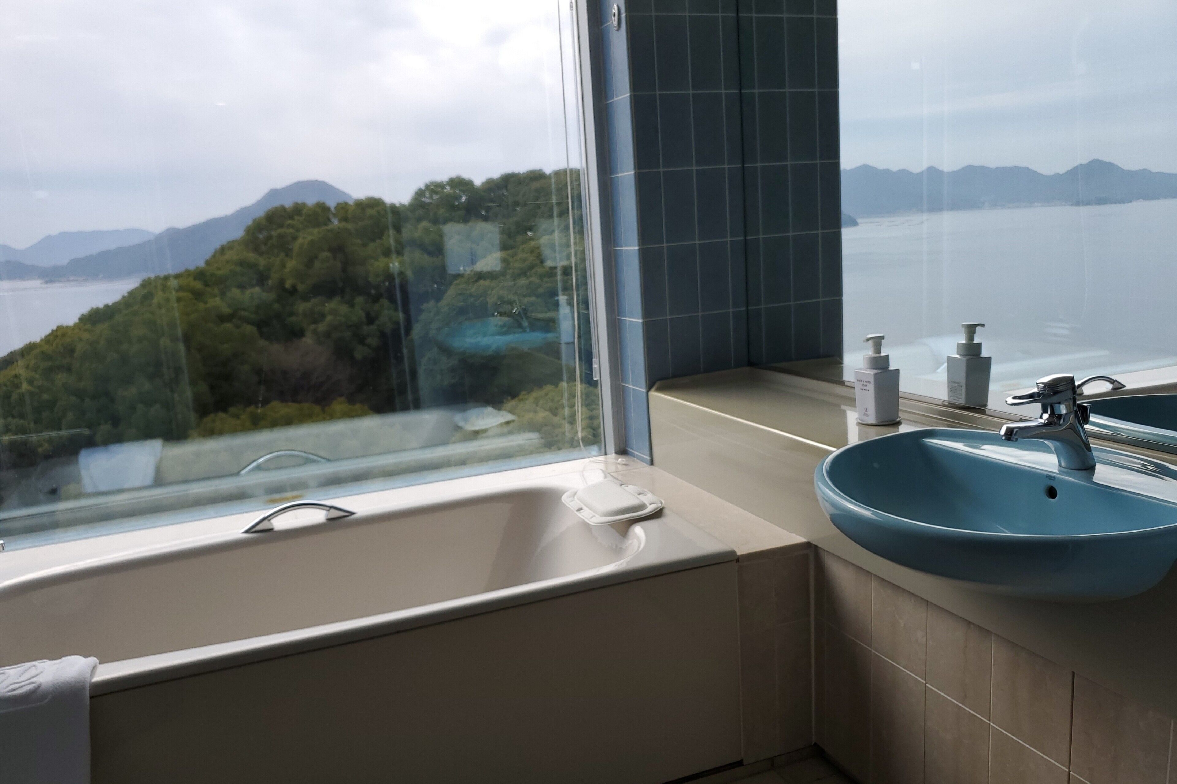 A bathroom with a bathtub and a sink, both with modern fixtures. A large window offers a view of a mountainous landscape and a body of water.
