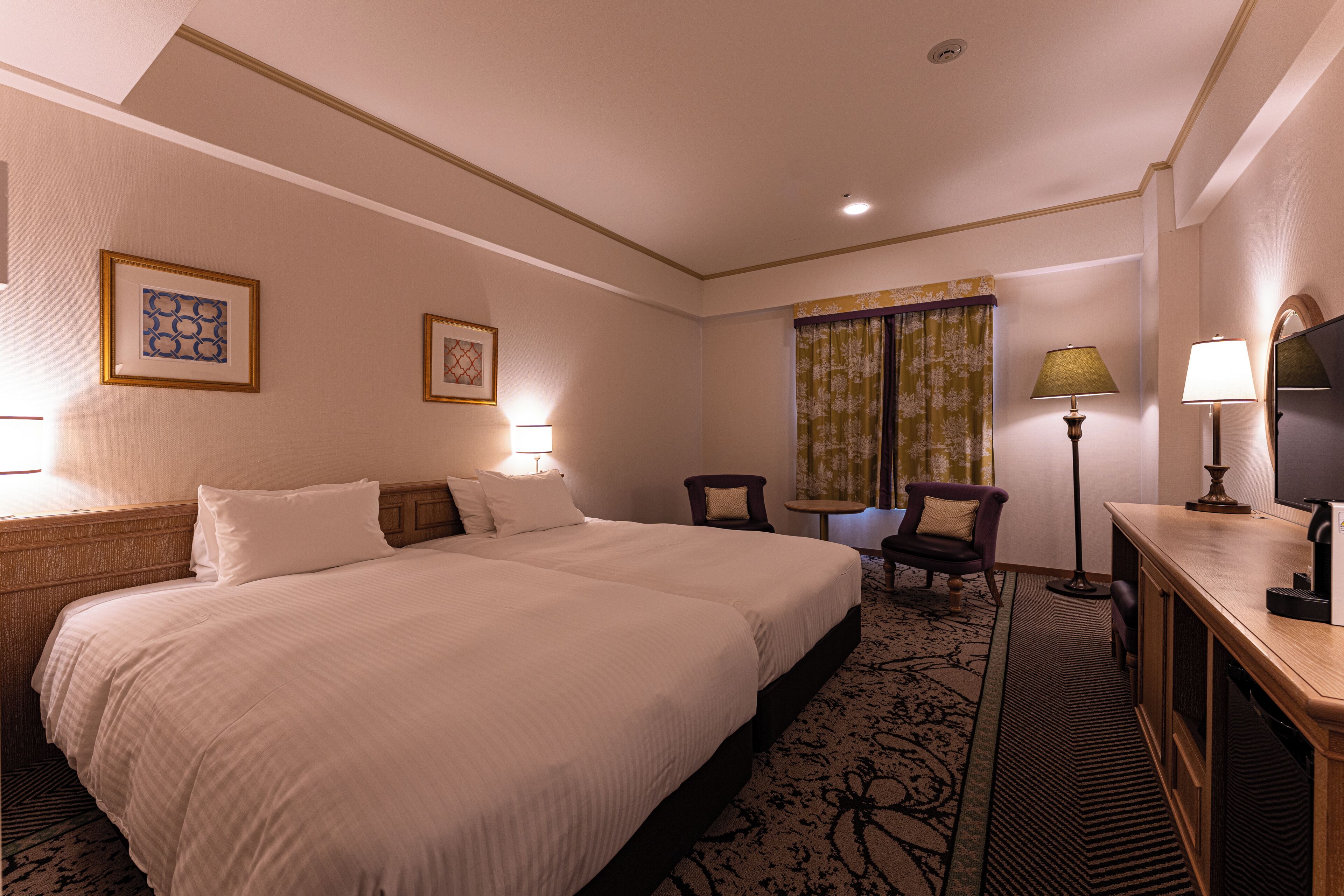 A hotel room with a large bed, a desk, a chair, a television, and two framed pictures on the wall.