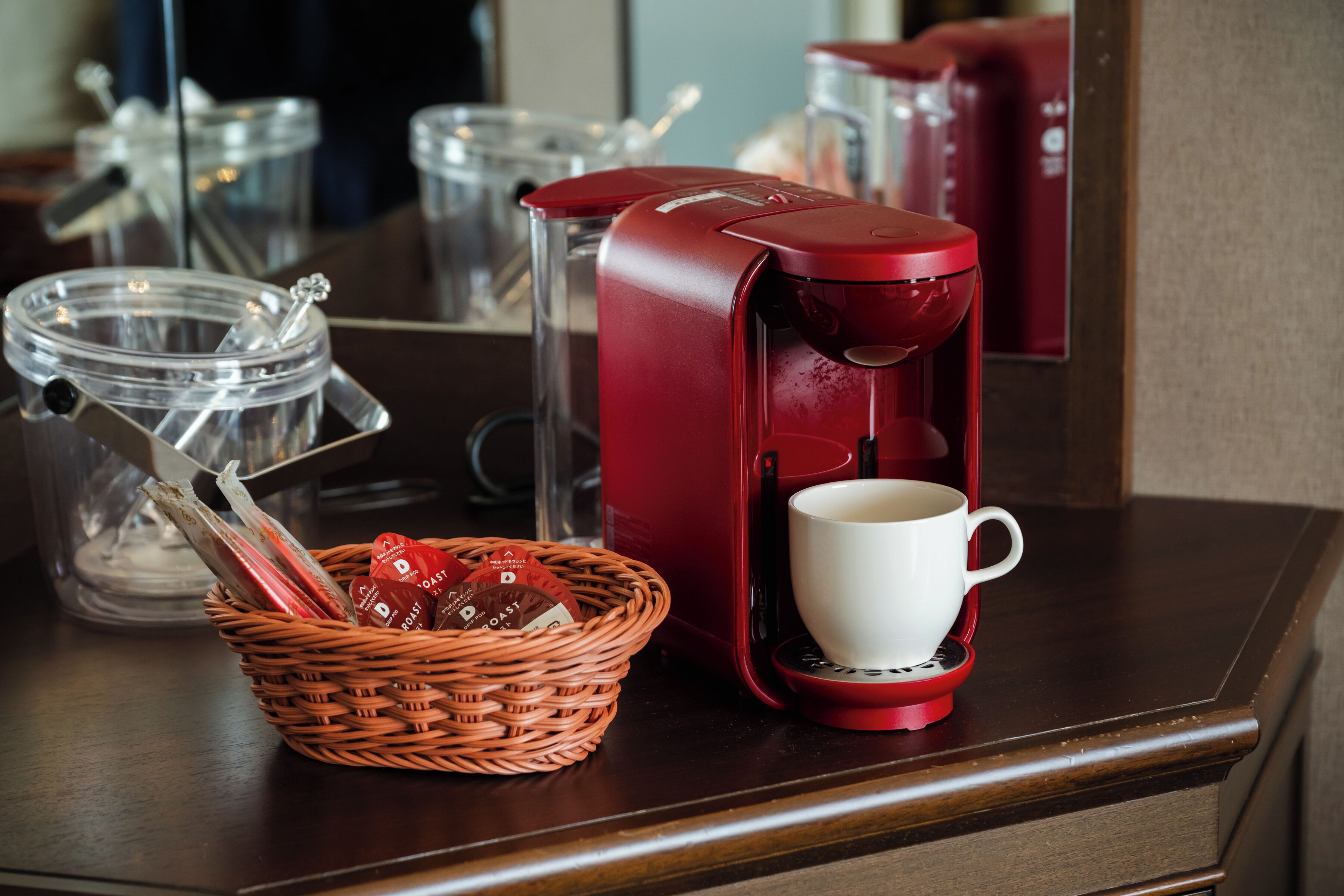 A red coffee maker with a white cup on a wooden surface, a wicker basket with coffee pods, and clear glass containers in the background.