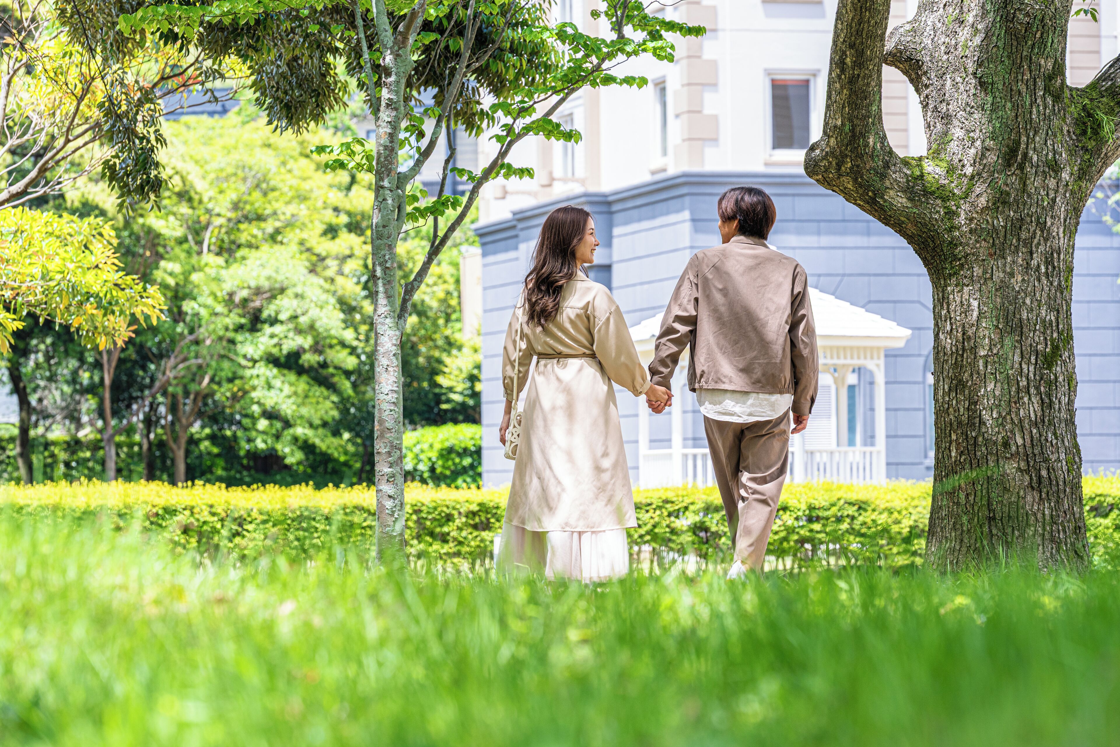 Una pareja caminando de la mano por un sendero cubierto de hierba, con árboles y un edificio al fondo.