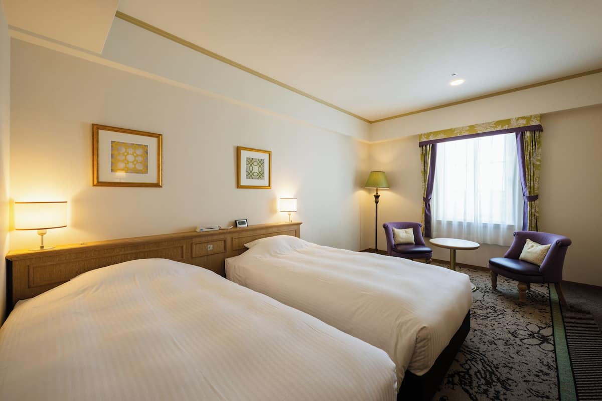 Ein Hotelzimmer mit zwei Betten, einem kleinen Tisch, zwei Stühlen, einer Lampe und gerahmten Bildern an der Wand.