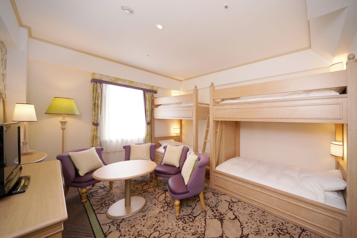 Ein Hotelzimmer mit Stockbetten, einem runden Tisch, violetten Sesseln, einem Fernseher und einem Fenster mit Vorhängen.