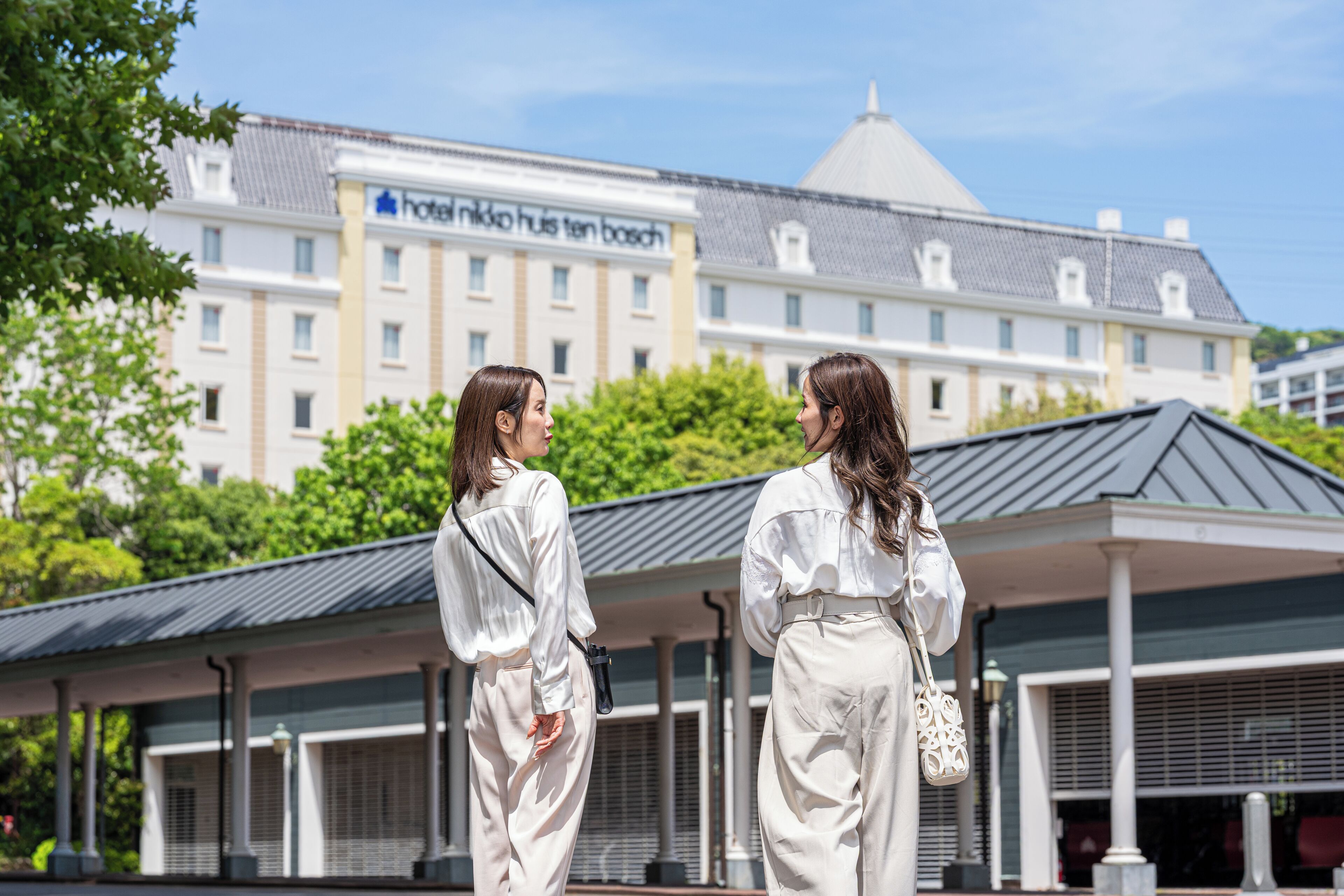 Two individuals standing in front of a hotel with a covered outdoor area and a building with a sign that reads 'Hotel ten bosch'.