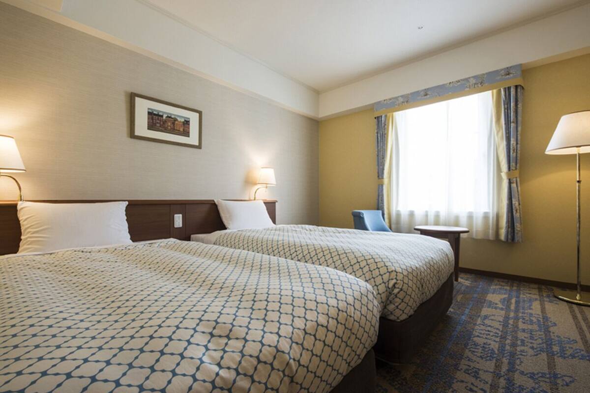 Ein Hotelzimmer mit zwei Betten, einem Schreibtisch, einem Stuhl, einer Lampe und einem fensterbepflanzten Fenster.