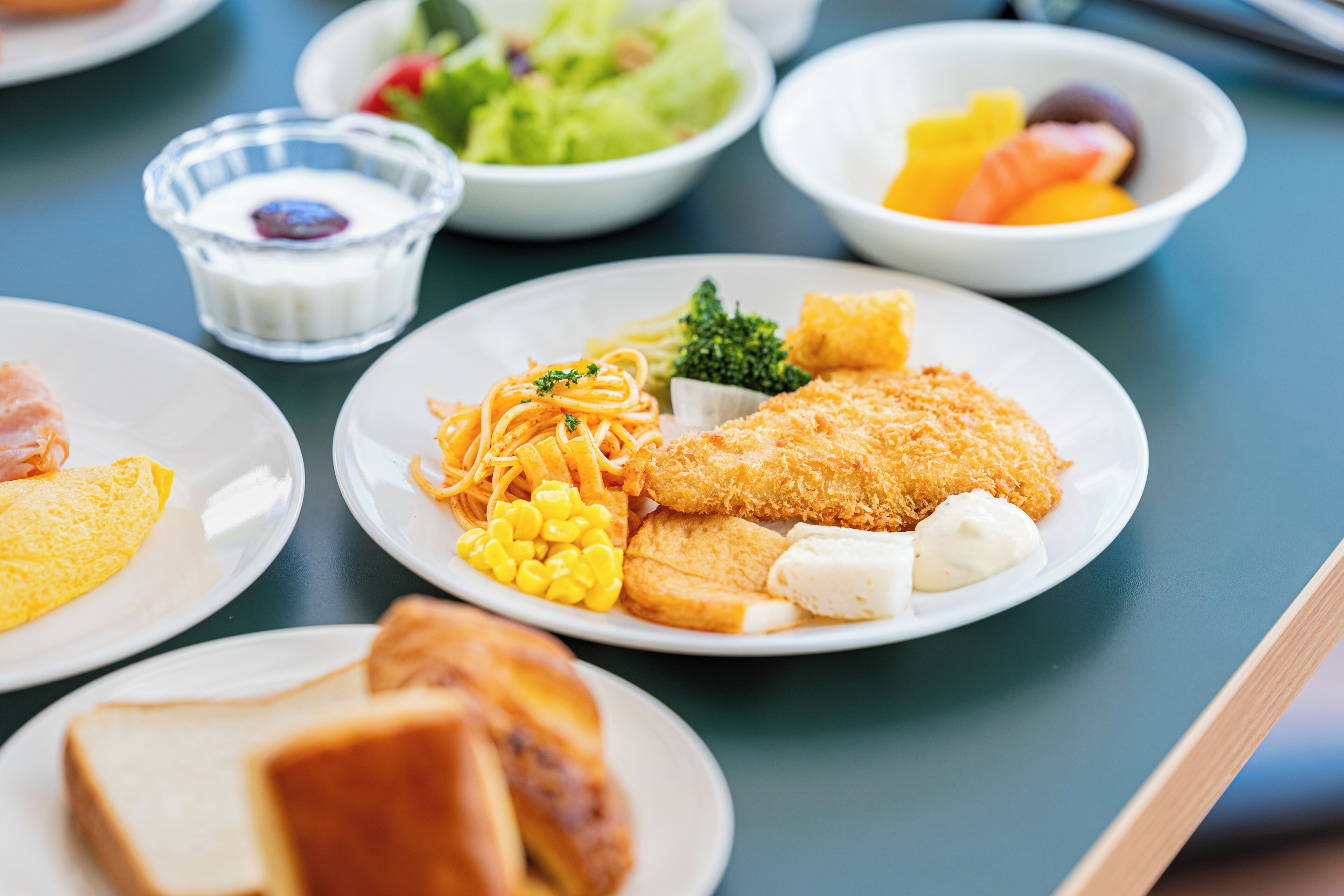 A table with various dishes including a breaded fish fillet, pasta, corn, and a salad.