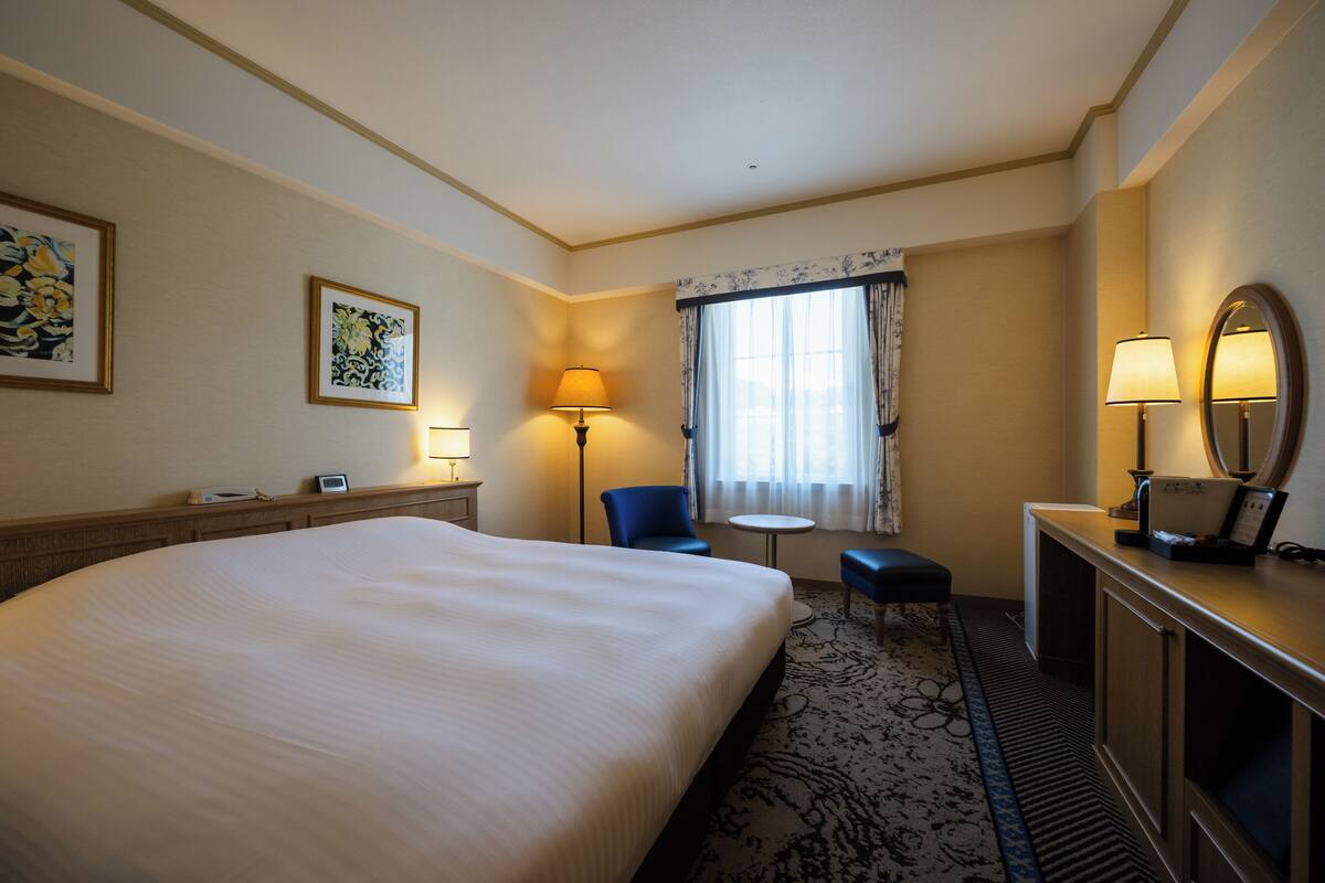 Ein Hotelzimmer mit einem großen Bett, einem Schreibtisch mit Spiegel, einem Sessel, einem kleinen Tisch und zwei Lampen.