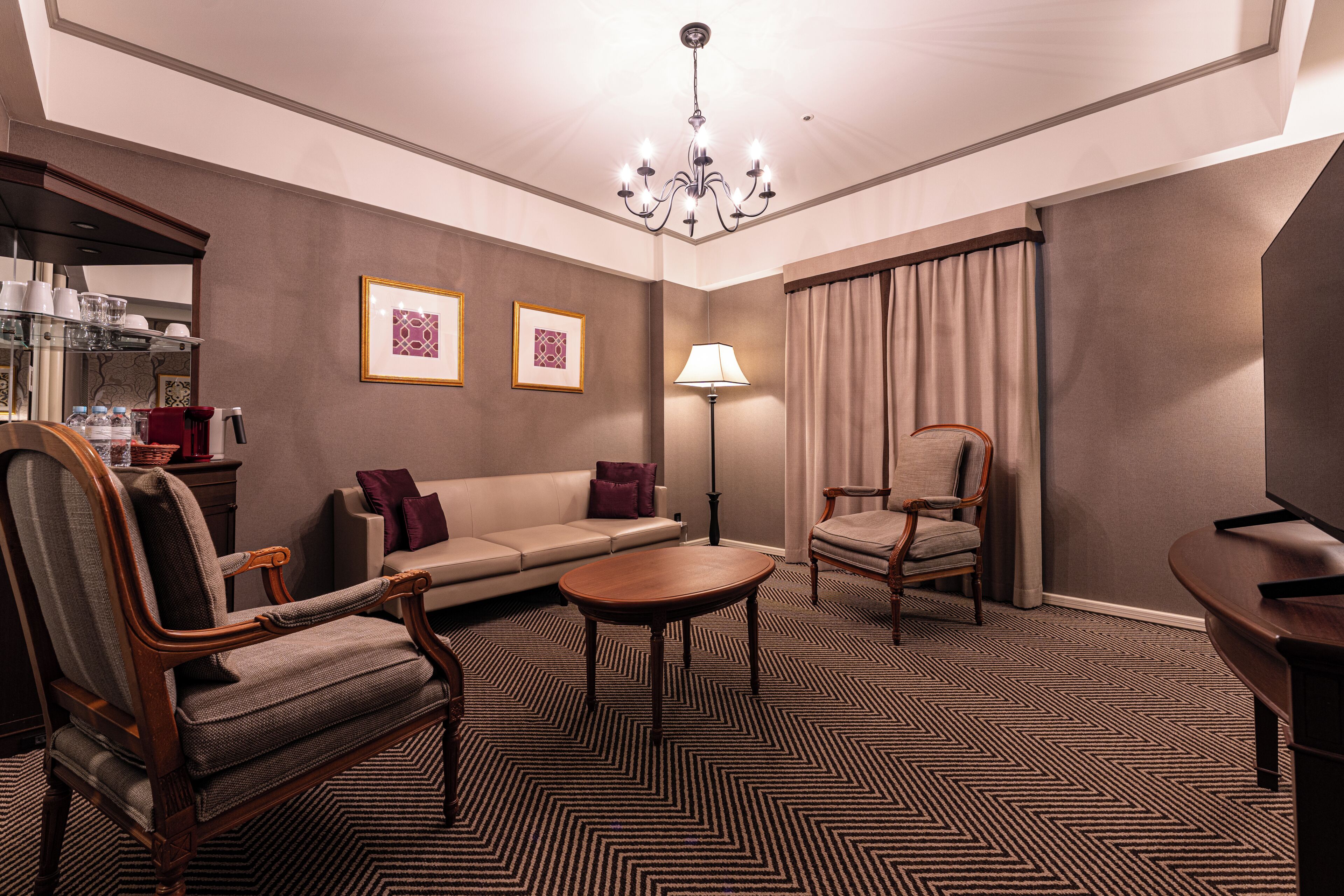 A hotel room with a sofa, two armchairs, a small table, a lamp, a chandelier, and a bed.