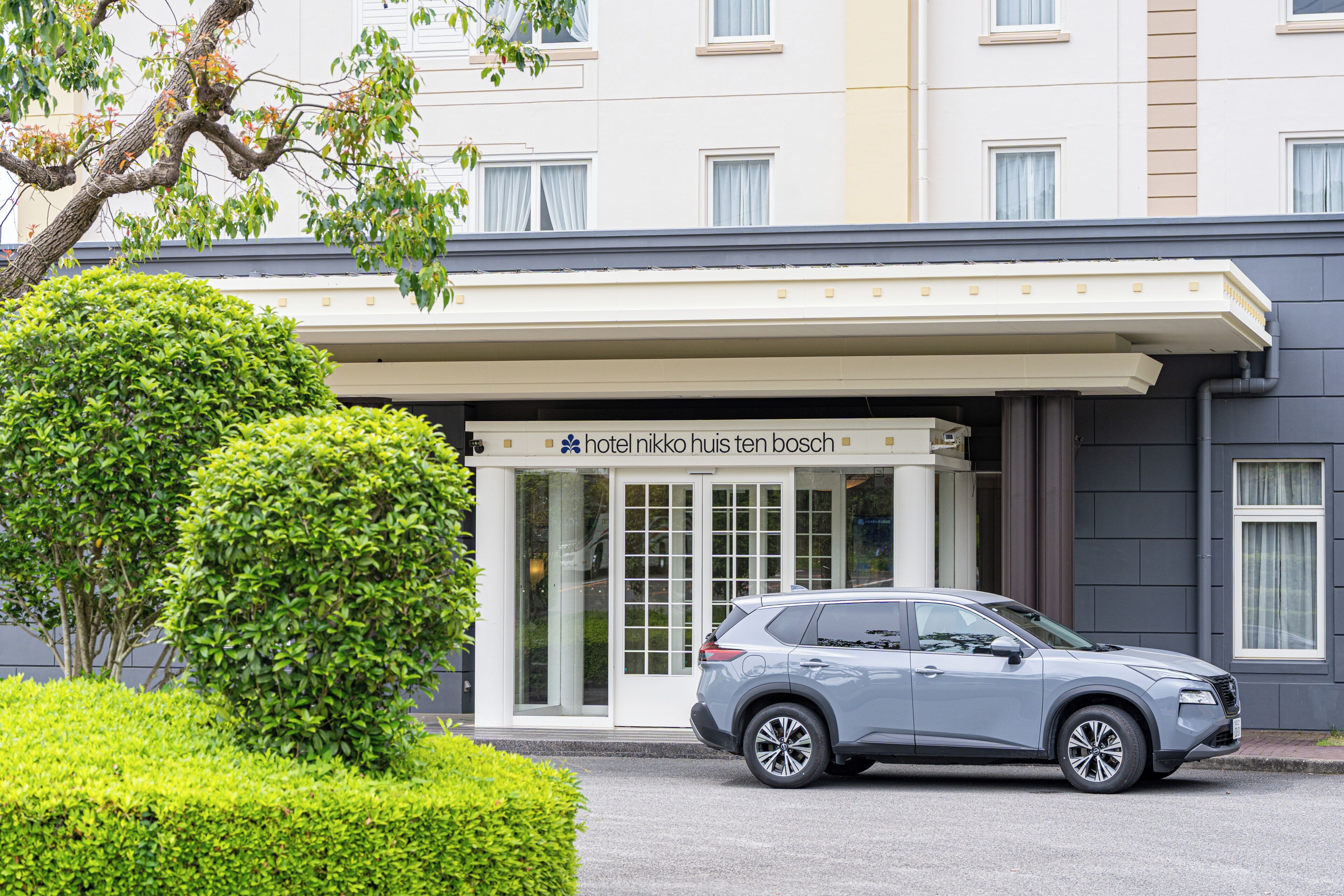 A modern hotel entrance with a clear sign, a well-maintained driveway, and a neatly trimmed hedge.