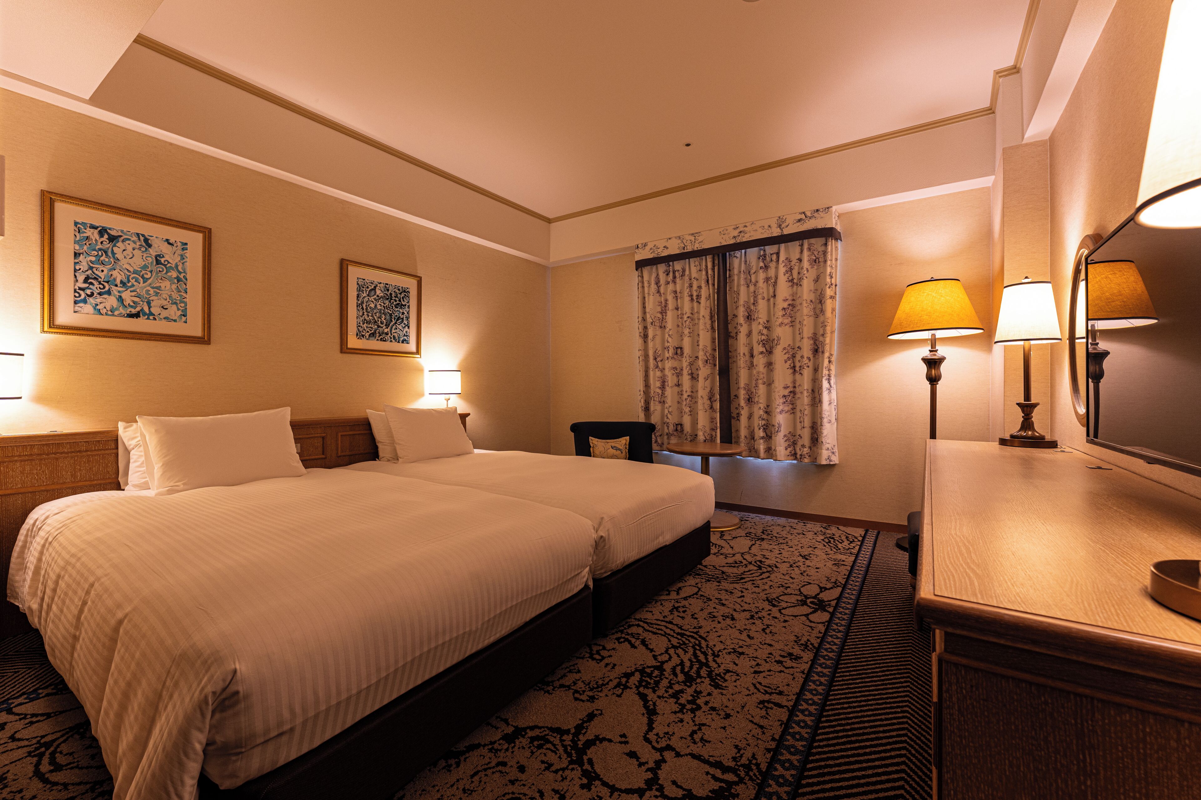 A hotel room with a double bed, two bedside tables with lamps, a small chair, a desk with a lamp, and a large mirror.