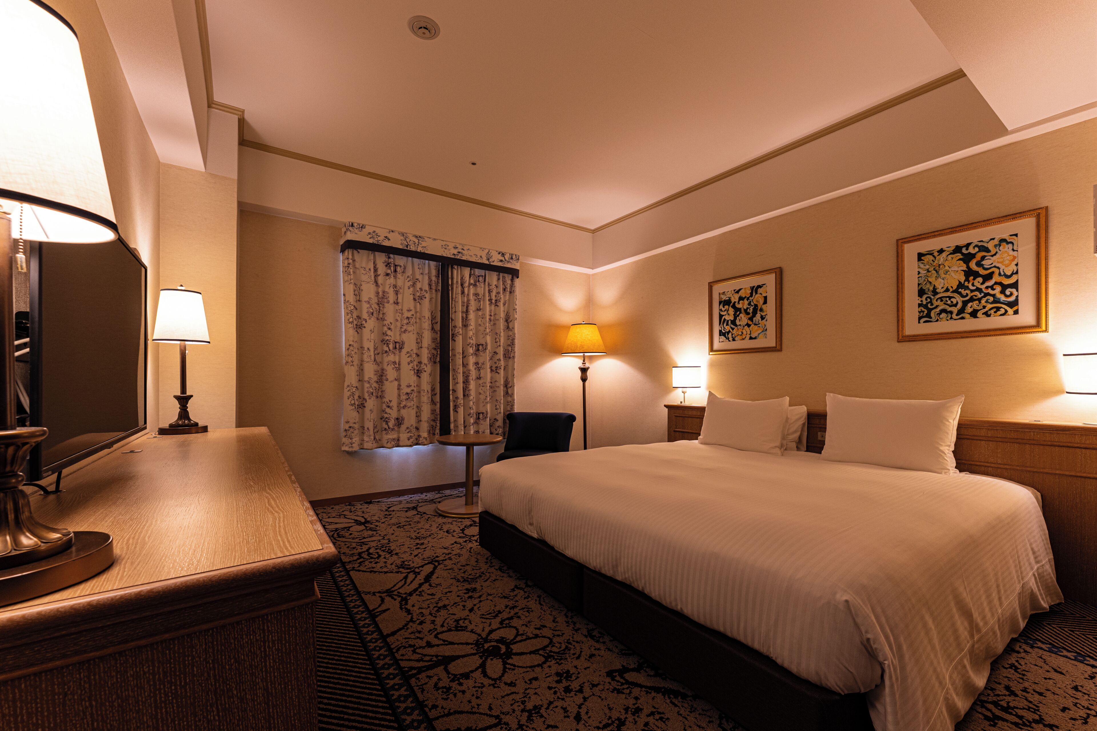 A hotel room with a bed, bedside lamps, a desk with a lamp, a chair, and a carpeted floor.