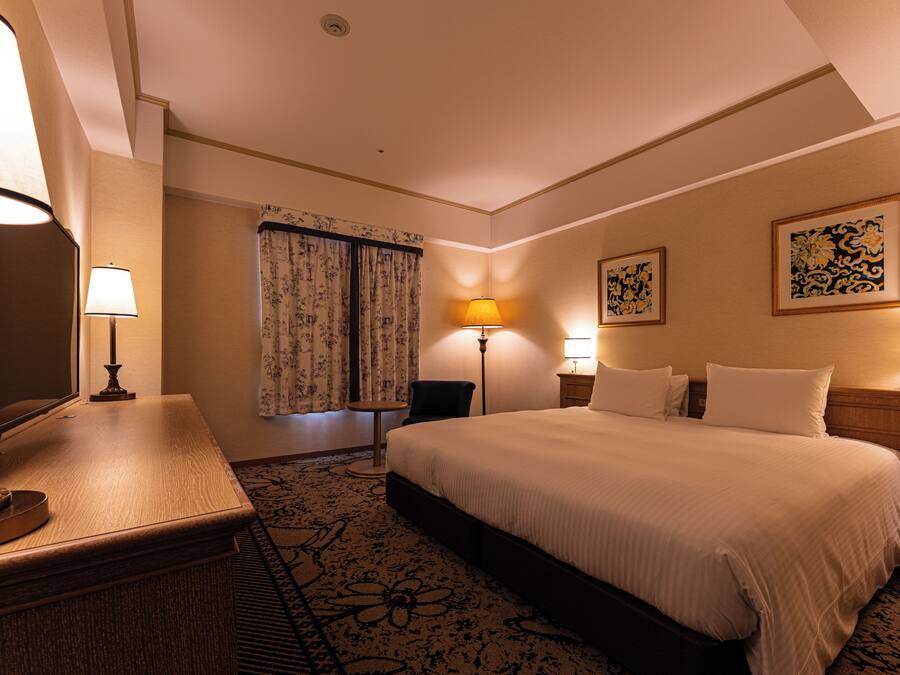 A hotel room with a bed, bedside lamps, a desk with a lamp, a chair, and a carpeted floor.