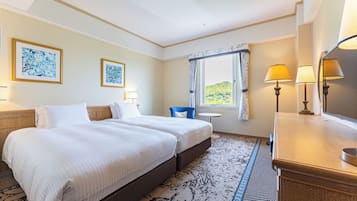 A hotel room with two beds, a small table, a chair, a desk with a lamp, and a large window with curtains.