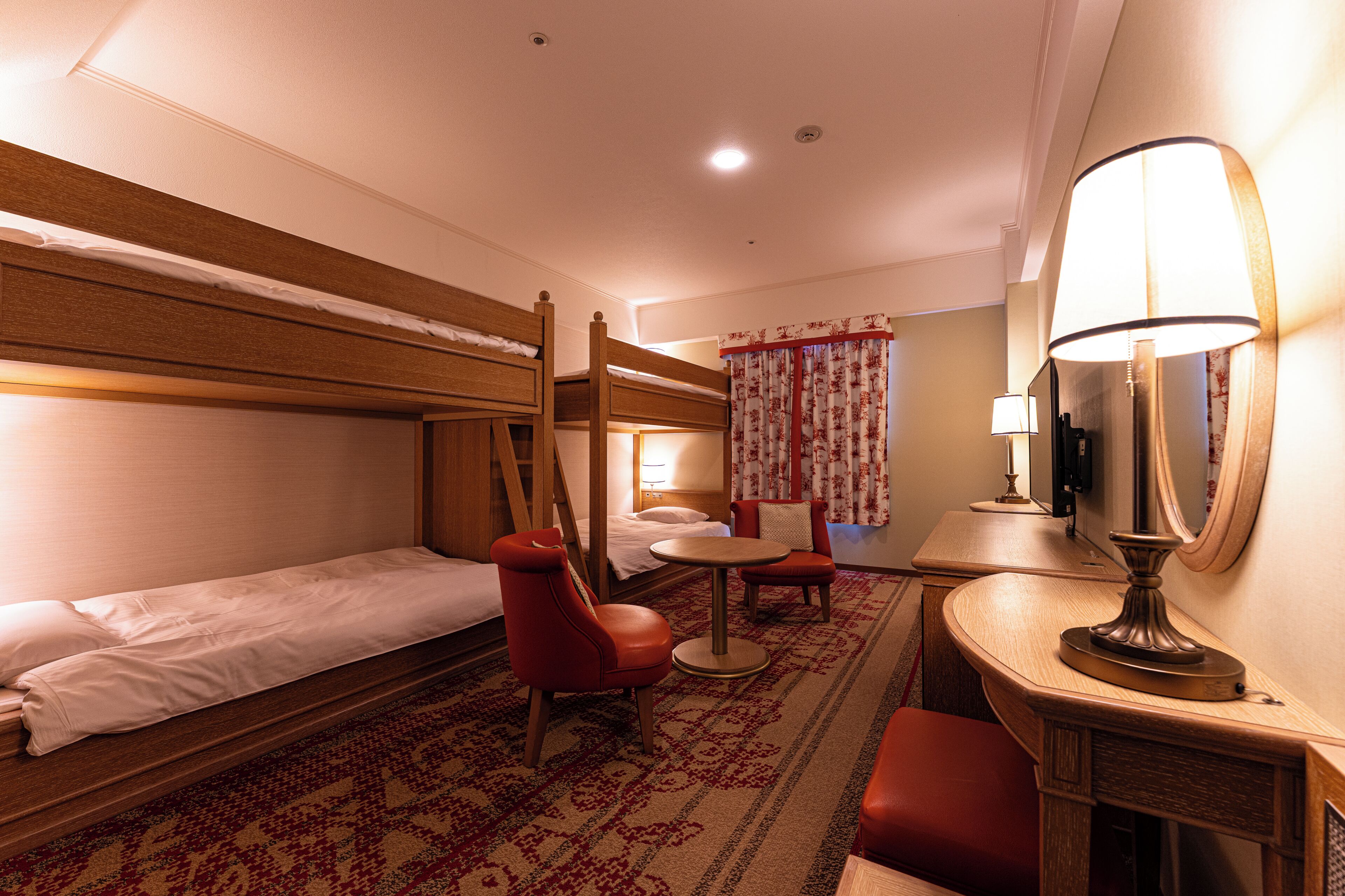 A hotel room with a bunk bed, a single bed, a desk with a lamp, a chair, and a small table.