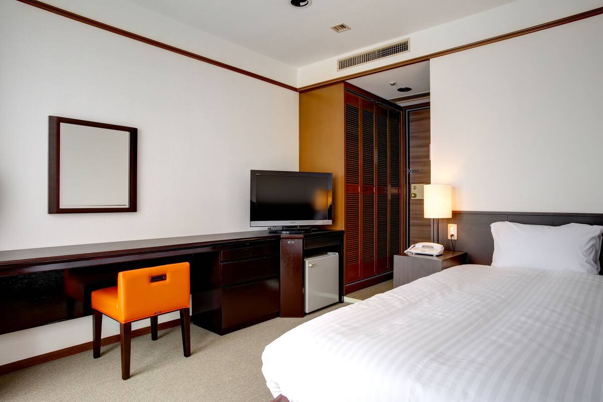 Ein Hotelzimmer mit einem Bett, einem Schreibtisch mit Fernseher, einem Sessel, einem kleinen Kühlschrank, einem Nachttisch mit Lampe und einem Kleiderschrank.