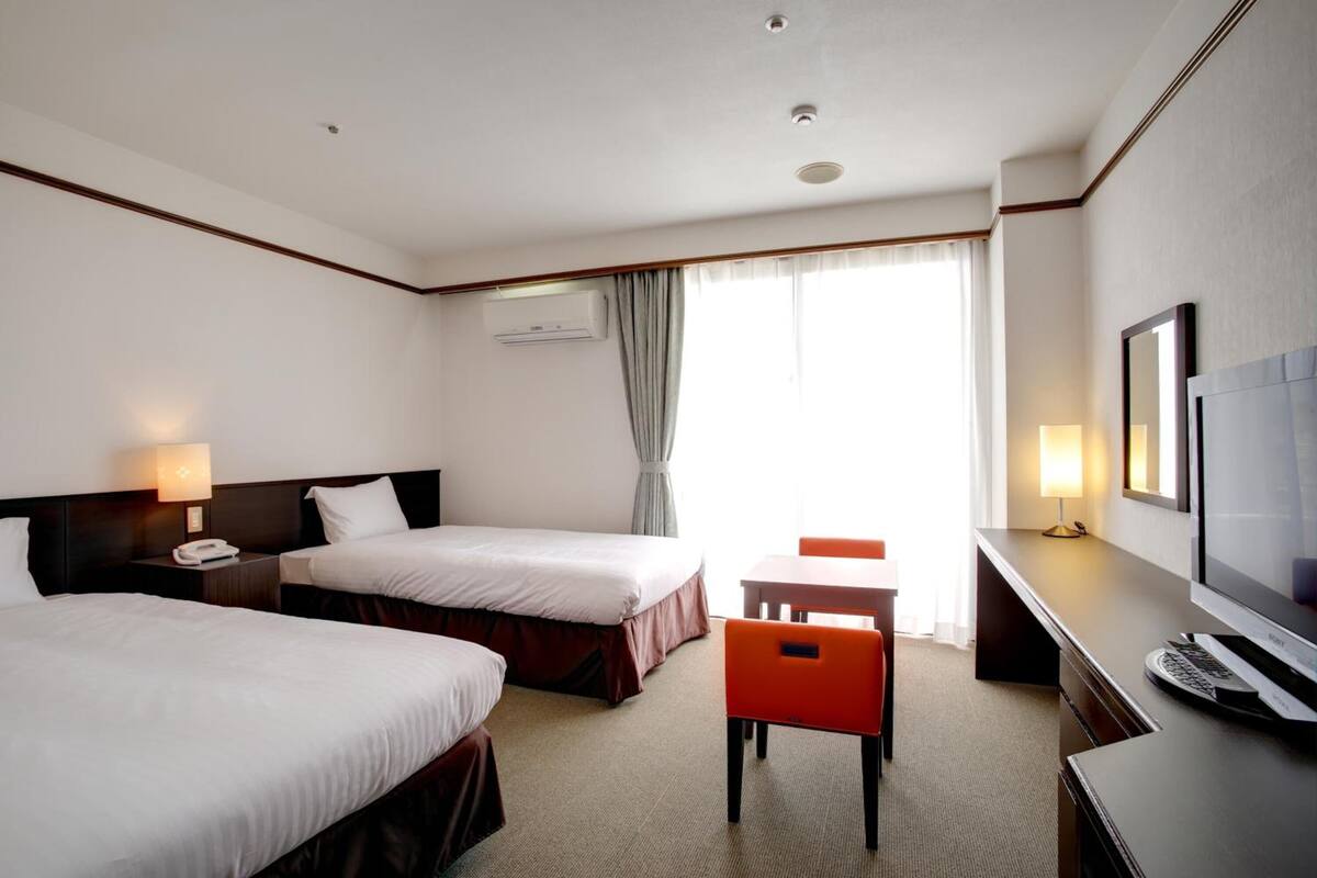 Ein Hotelzimmer mit zwei Betten, einem Schreibtisch, einem Stuhl, einem Fernseher und einem Fenster mit Vorhängen.